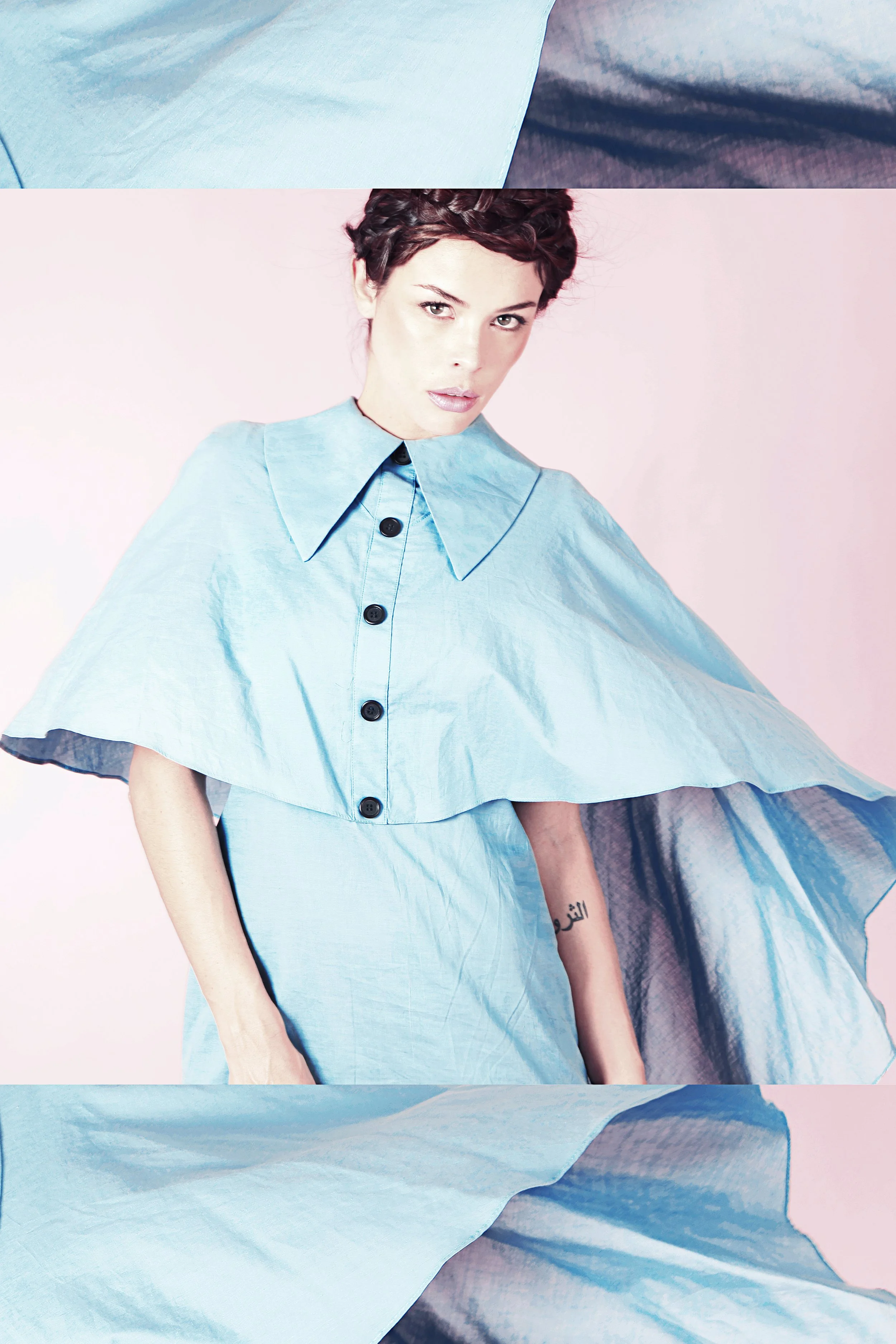 Close-up of a woman with braided hair wearing a light blue button-up shawl, sitting against a pastel pink background with fabric draped on her side.