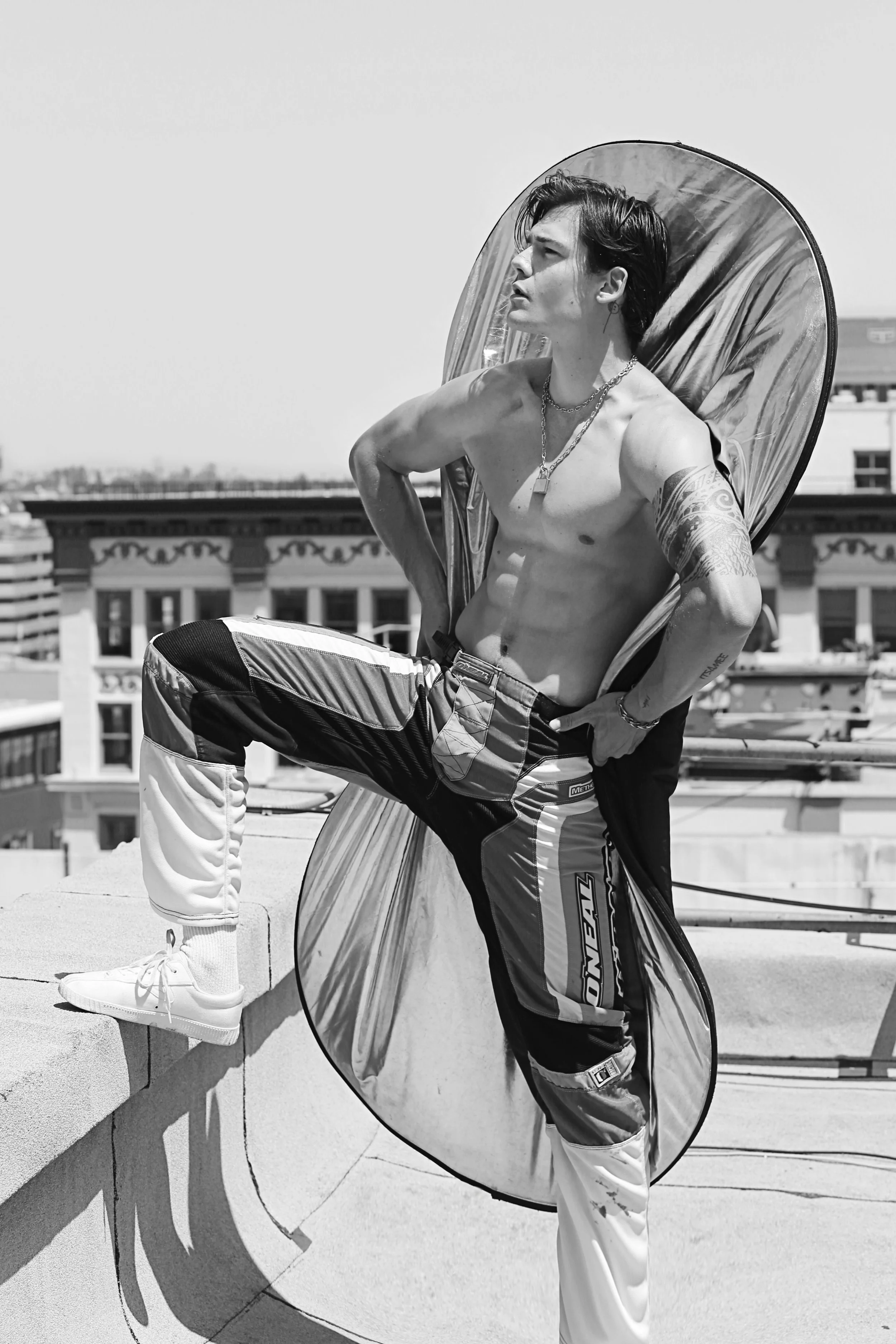 A shirtless young man with tattoos and jewelry, sitting on a rooftop, with a reflector propped behind him, city buildings in the background, black and white photograph.