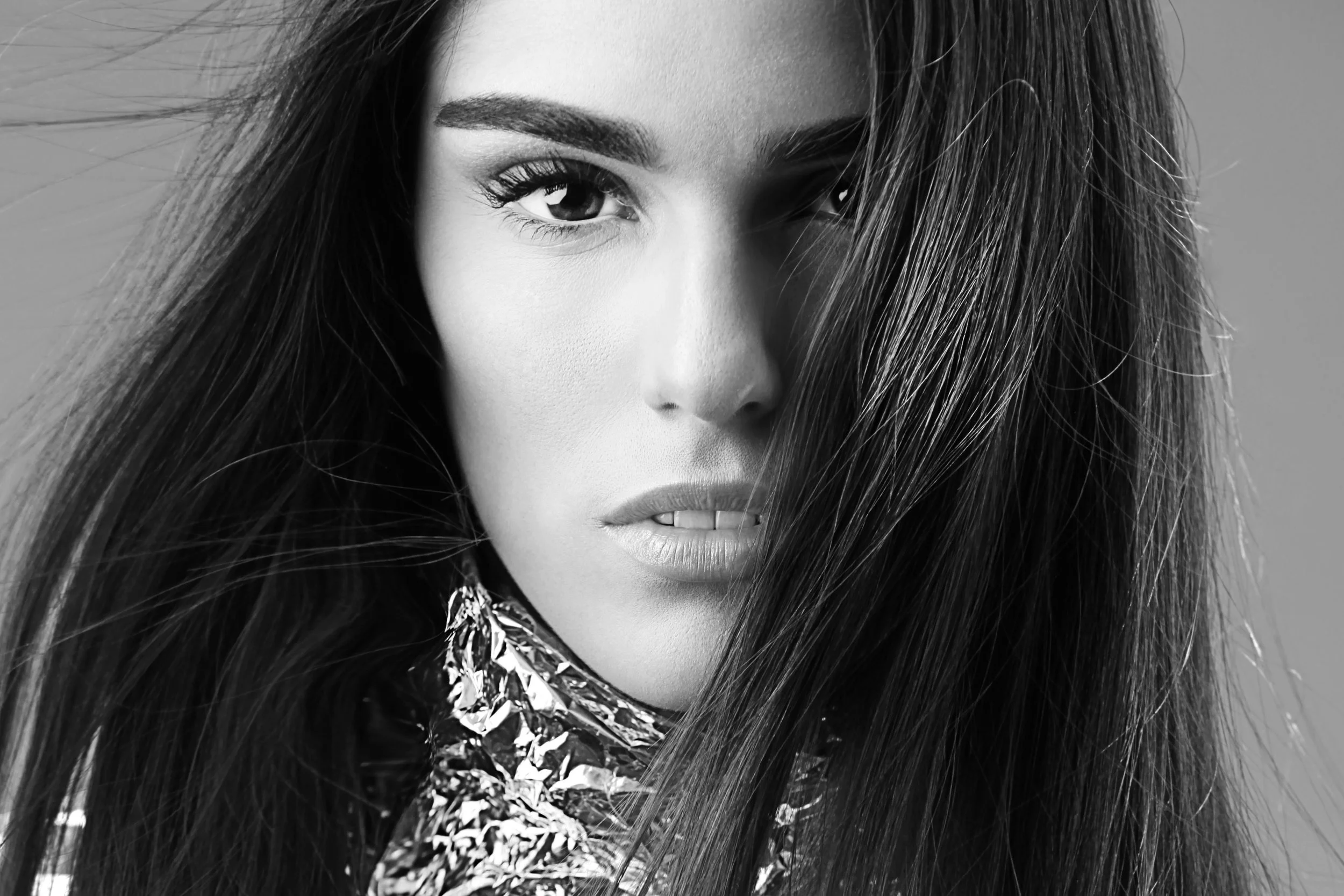 Black and white close-up portrait of a woman with long hair covering part of her face, wearing a shiny, textured garment.