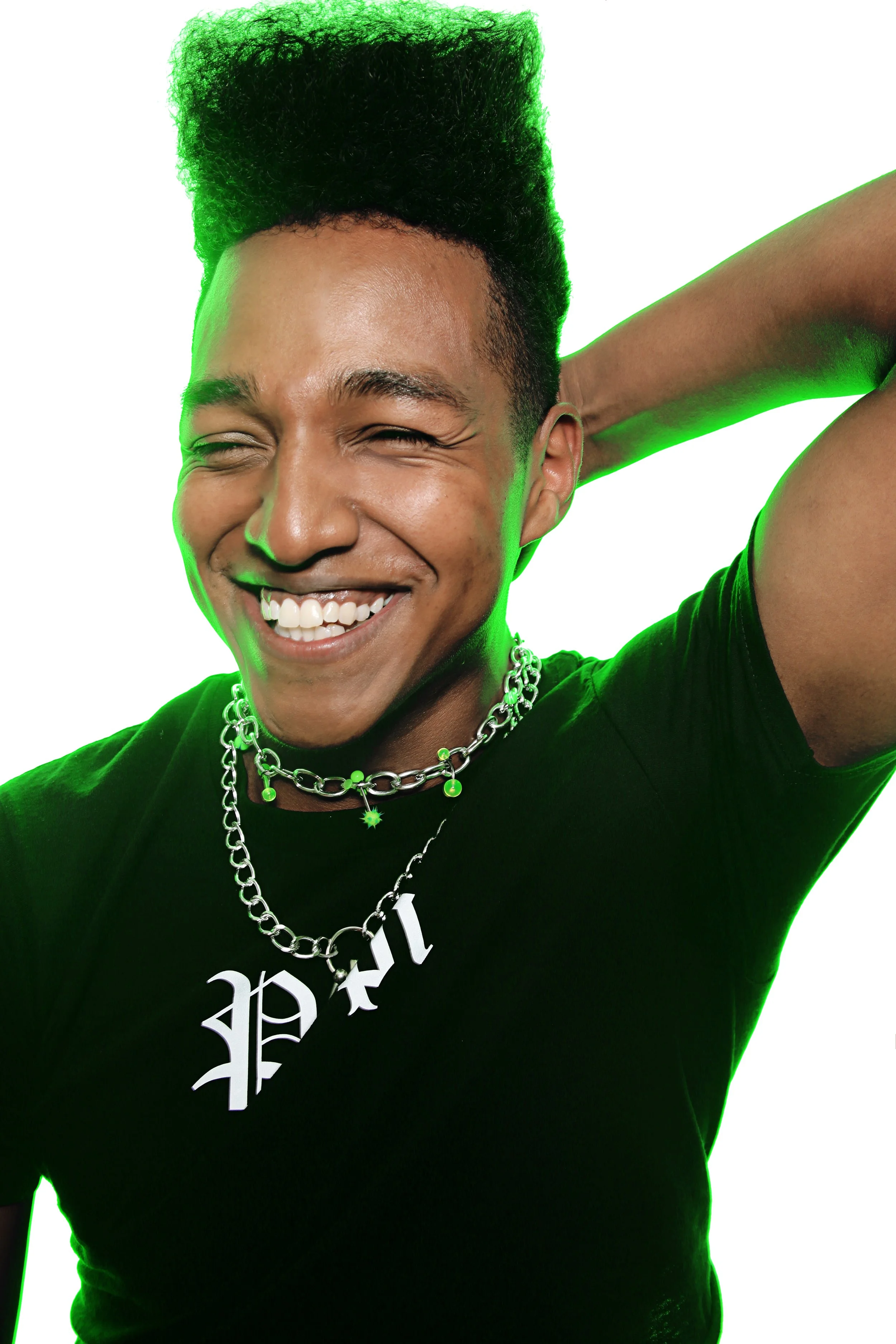 A person with a big smile, wearing a black shirt with white lettering, silver chain necklaces, and raising an arm behind their head against a white background with a green glow outline.