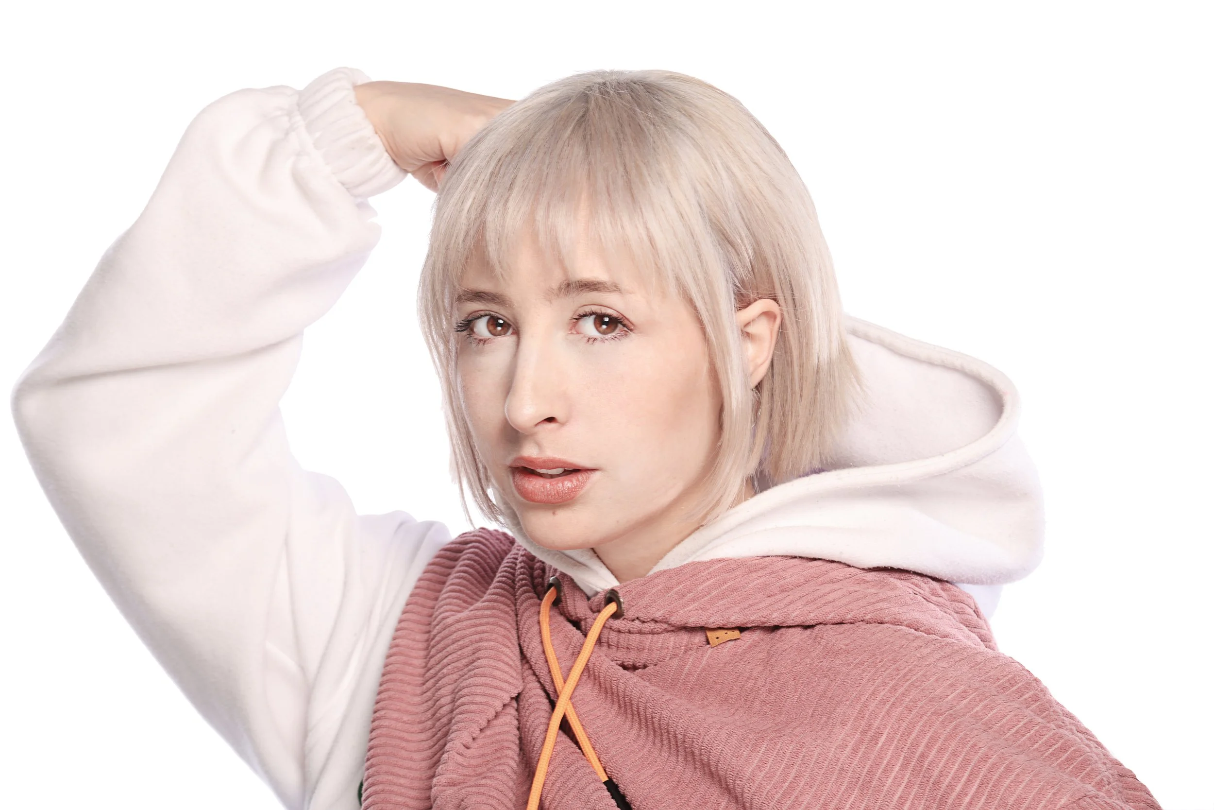 A young woman with platinum blonde hair, wearing a white hoodie and rust-colored pants, is scratching her head and looking directly at the camera against a plain white background.