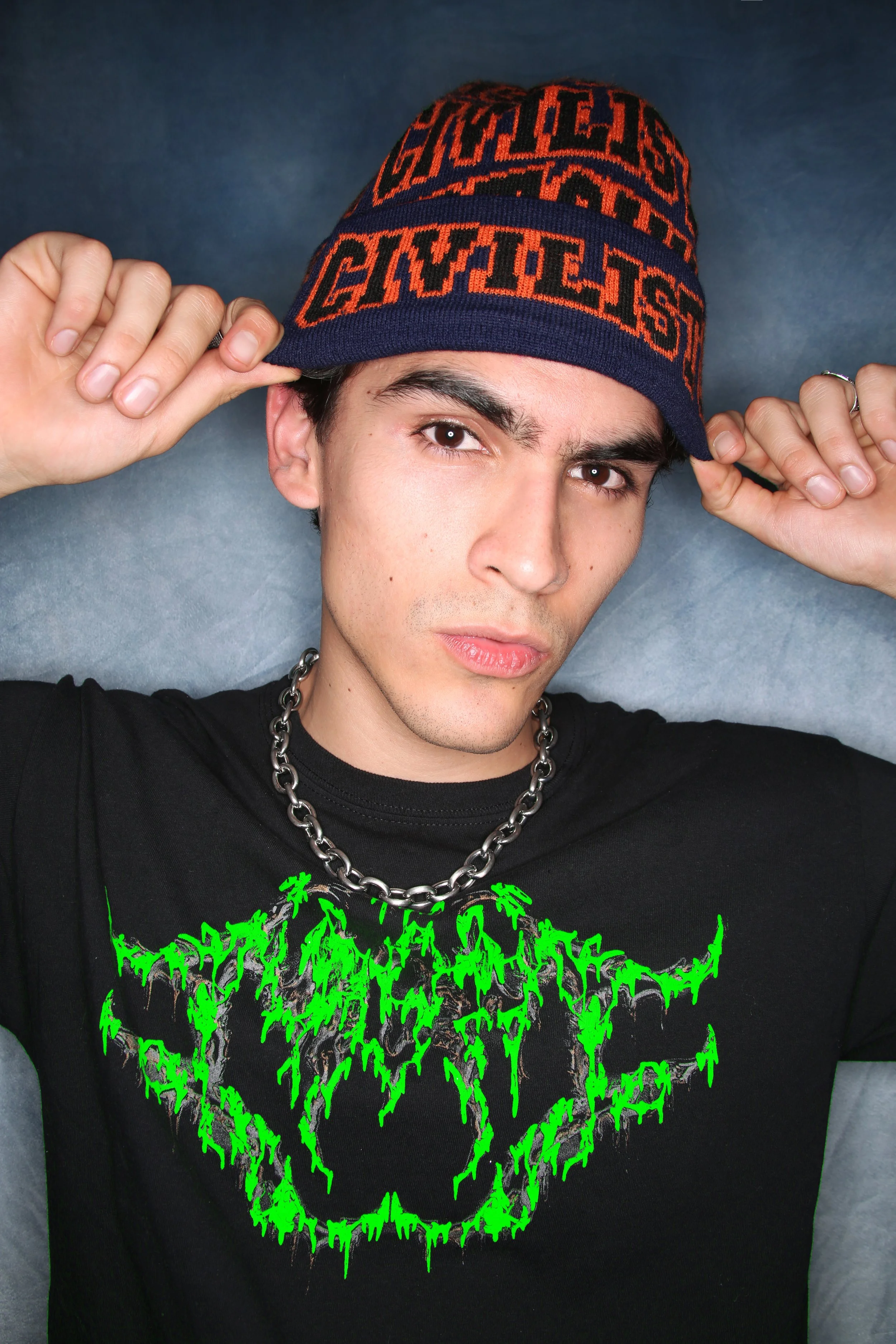 A young man with dark hair and light skin wearing a black T-shirt with bright green graphic designs and a chunky silver chain necklace, holding a navy blue and orange patterned beanie hat with text, against a gray background.