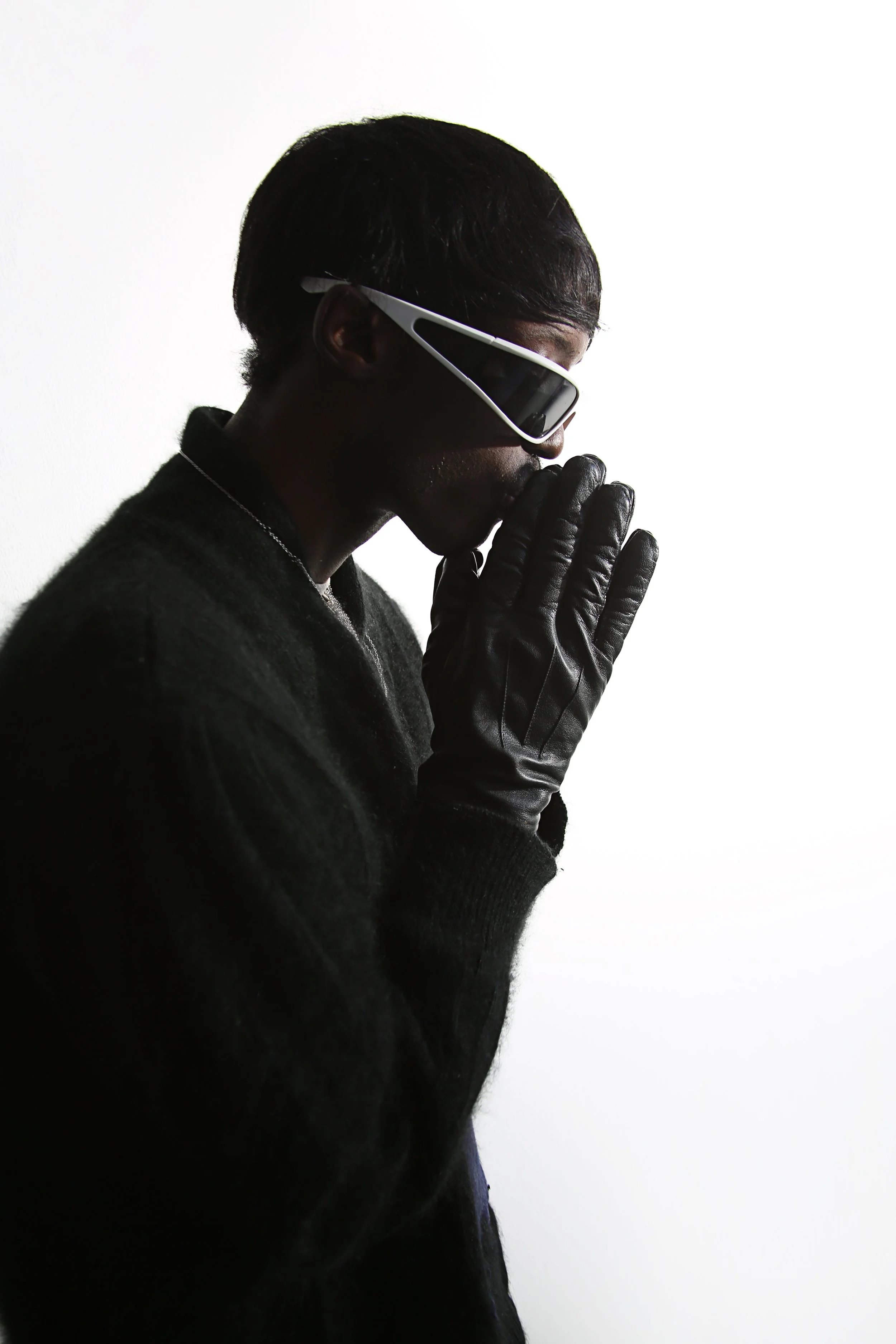 A person with short black hair wearing black gloves, a black sweater, and futuristic glasses, standing against a plain white background, with their hands pressed together near their face in a prayer-like pose.