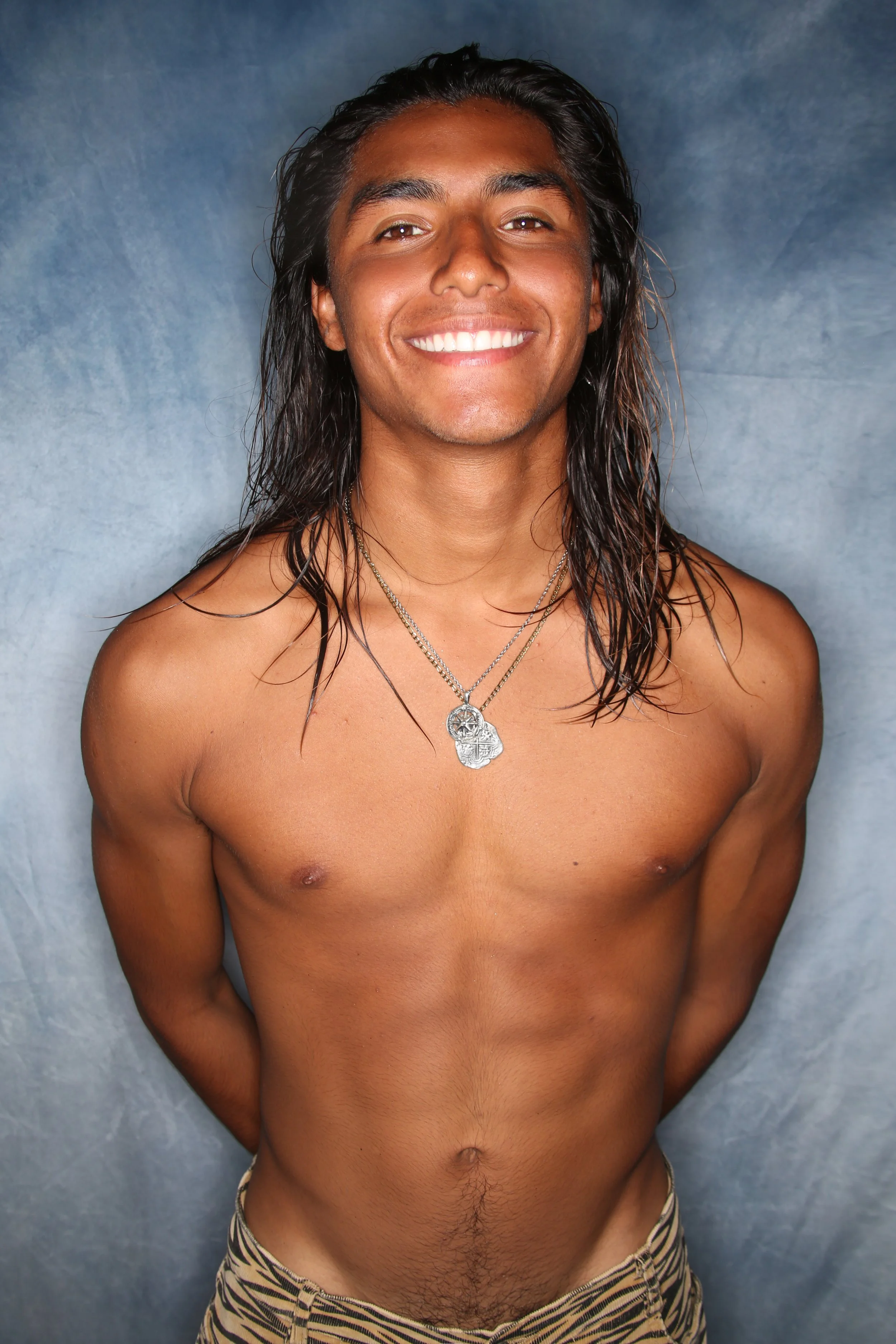 A shirtless young man with long wet hair smiling, wearing layered necklaces and tiger-striped shorts, standing against a blue-gray background.