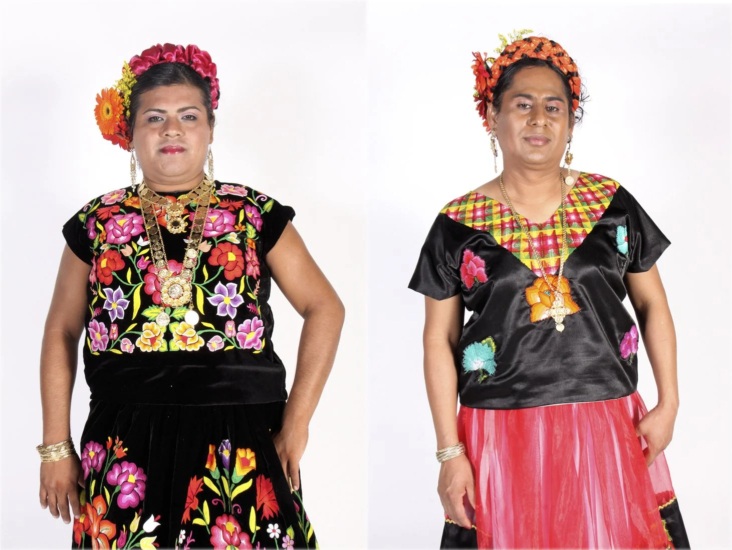 Two women dressed in traditional Mexican clothing, wearing colorful embroidered blouses, skirts, and floral headpieces, standing against a white background.