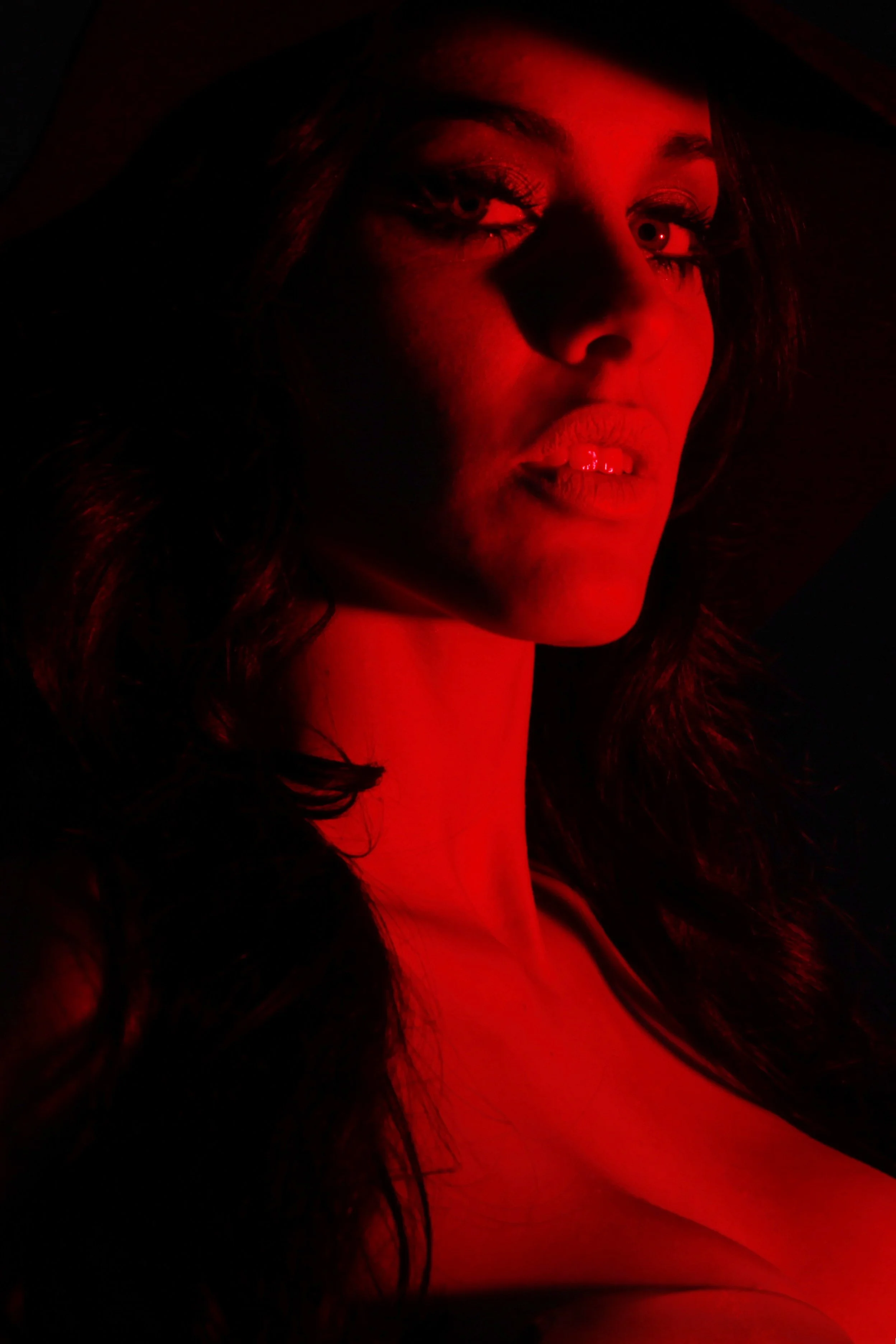 Close-up of a woman's face illuminated by red light, with long dark hair, makeup, and partially visible facial expression.