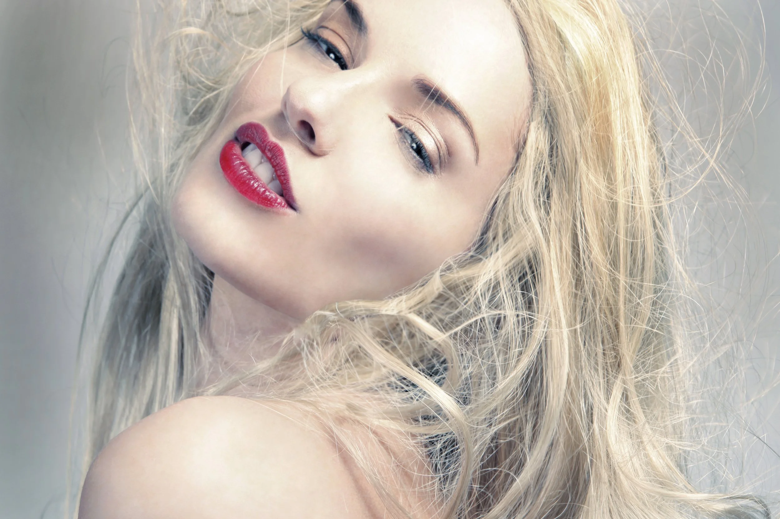 Close-up of a woman with blonde hair, makeup including red lipstick, wearing a playful expression with her lips slightly parted.