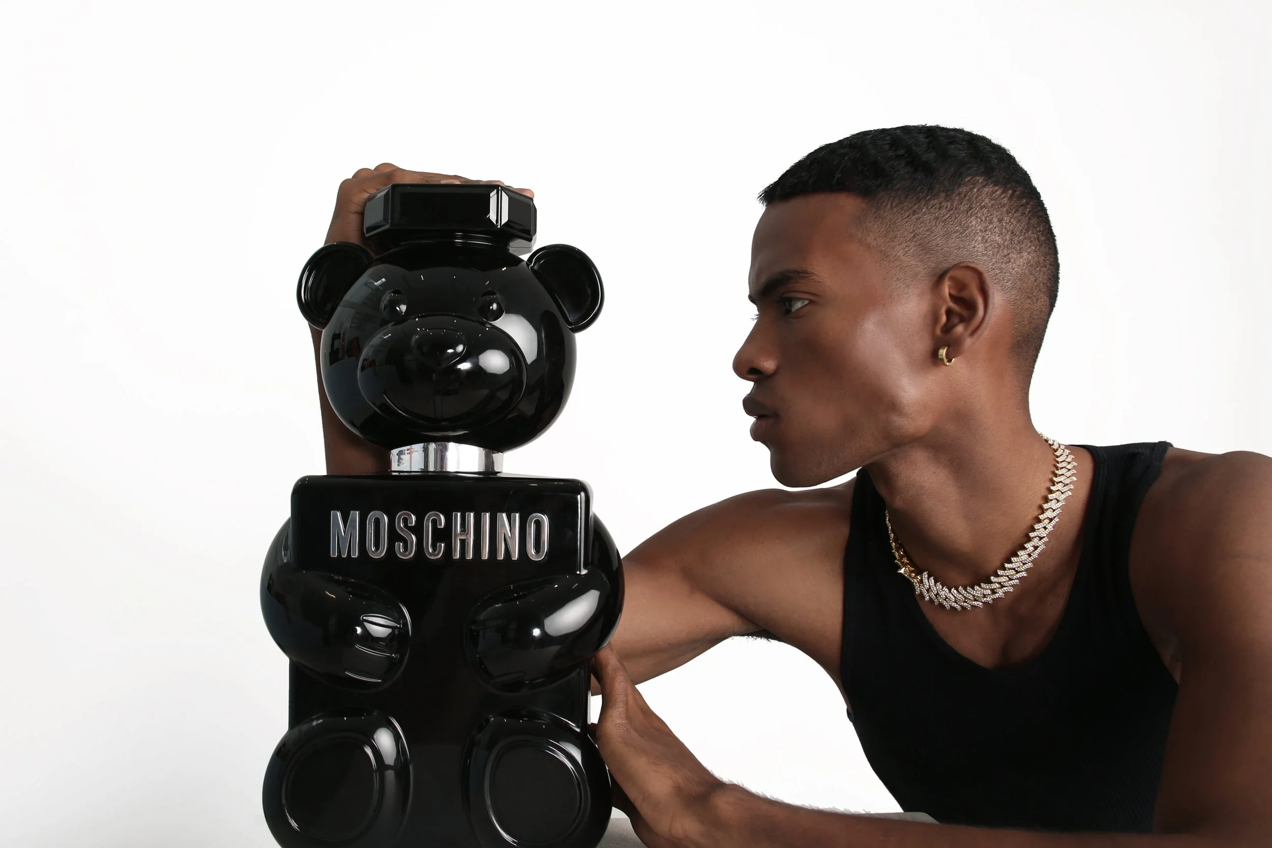 A man with short black hair, wearing a black sleeveless top, gold earrings, and a gold chain, looks at a black Moschino teddy bear-shaped speaker. The speaker has a shiny black finish and is labeled 'Moschino.'