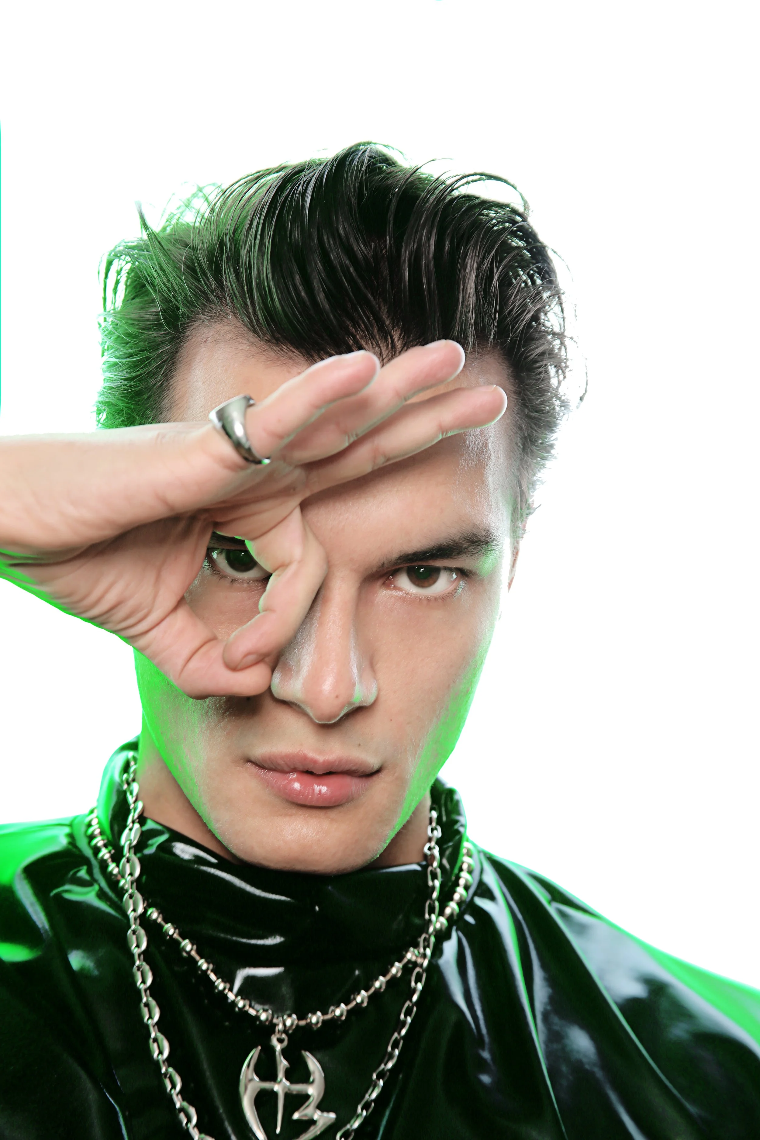 A person with short, styled dark hair making a lens flare with their hand, wearing a shiny black outfit and layered silver necklaces, against a bright white background.