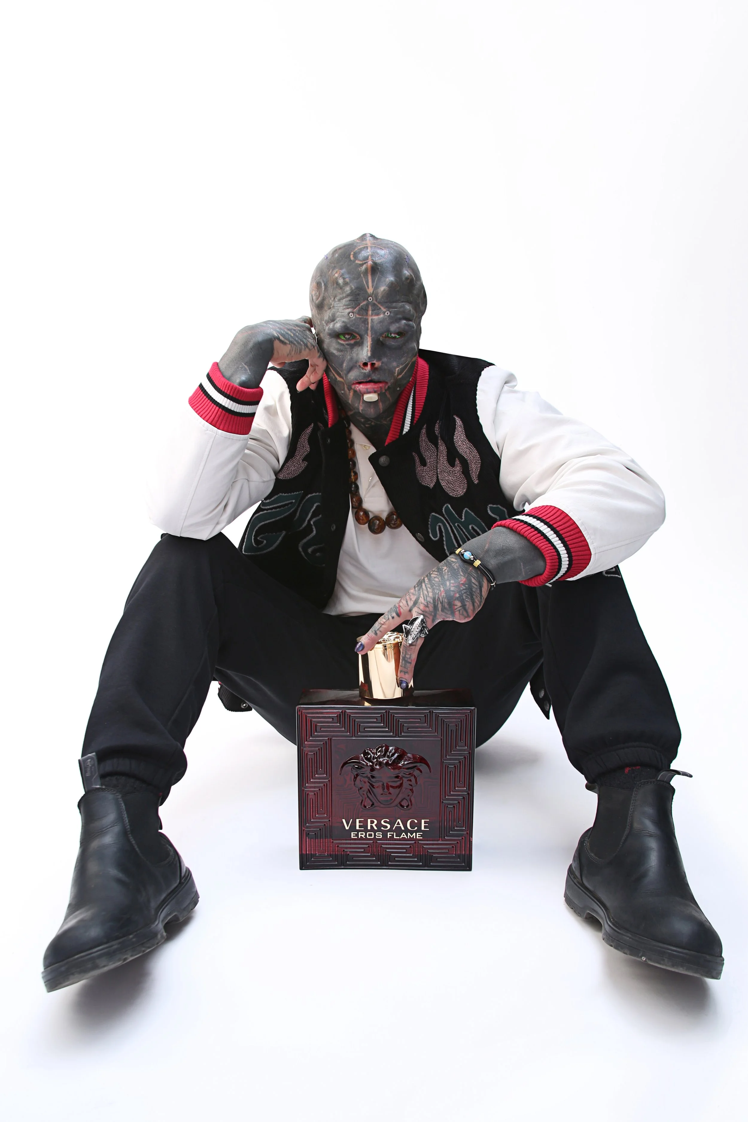 Person wearing a black and white varsity jacket, black boots, and a full face mask with tattoos, sitting with legs spread, holding a Versace Eros Flame perfume box between legs, against a plain white background.