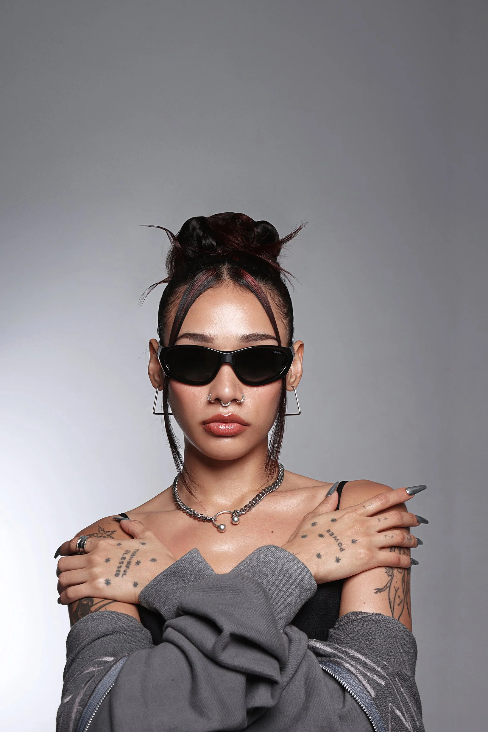 A young woman with short tattoos on her arms, wearing dark sunglasses, a grey off-the-shoulder top, silver jewelry, and long grey sleeves, with her arms crossed in front of her and fingers pointed upwards.