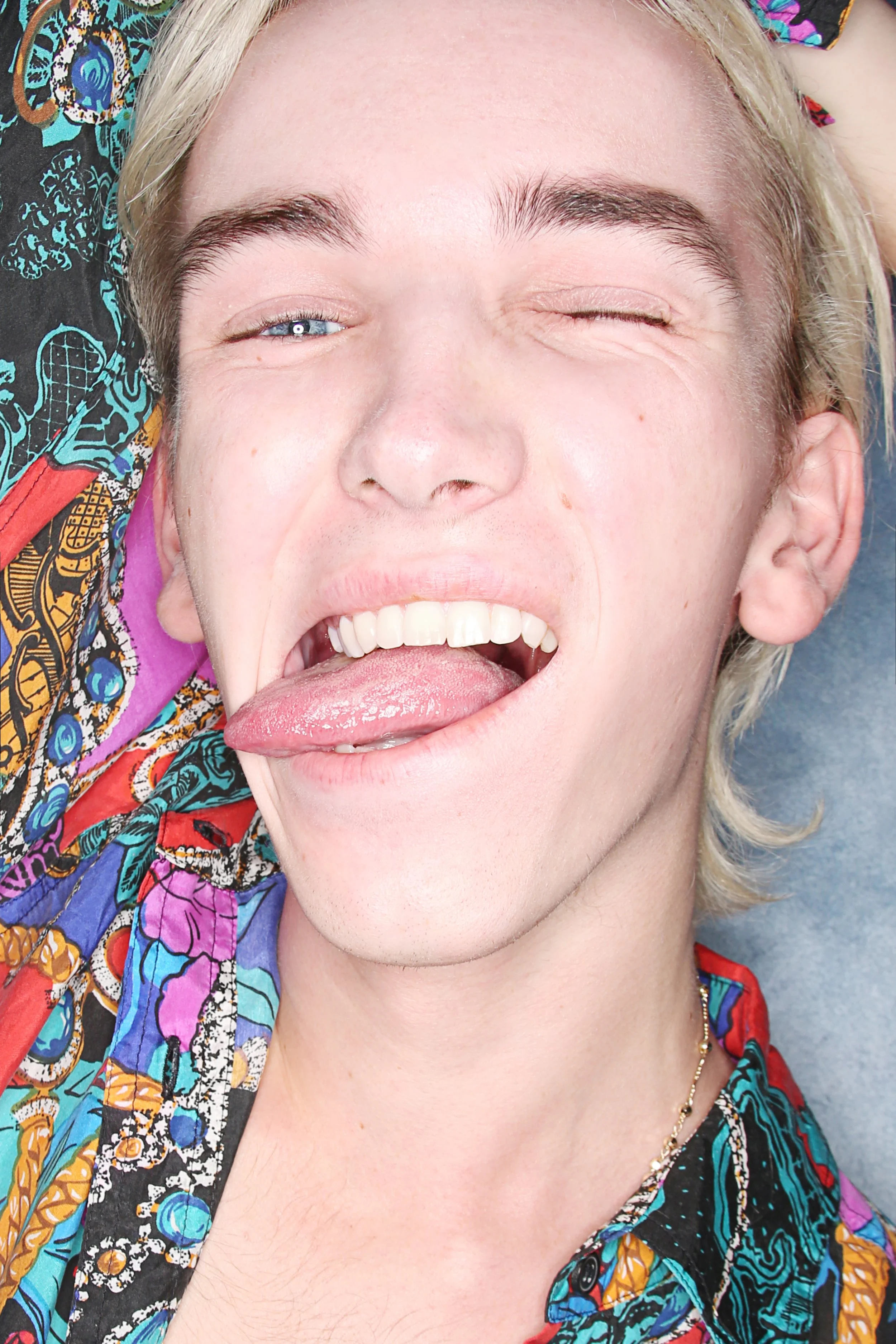 A close-up of a person with short blond hair and light skin, smiling with their tongue out, wearing a colorful patterned shirt.