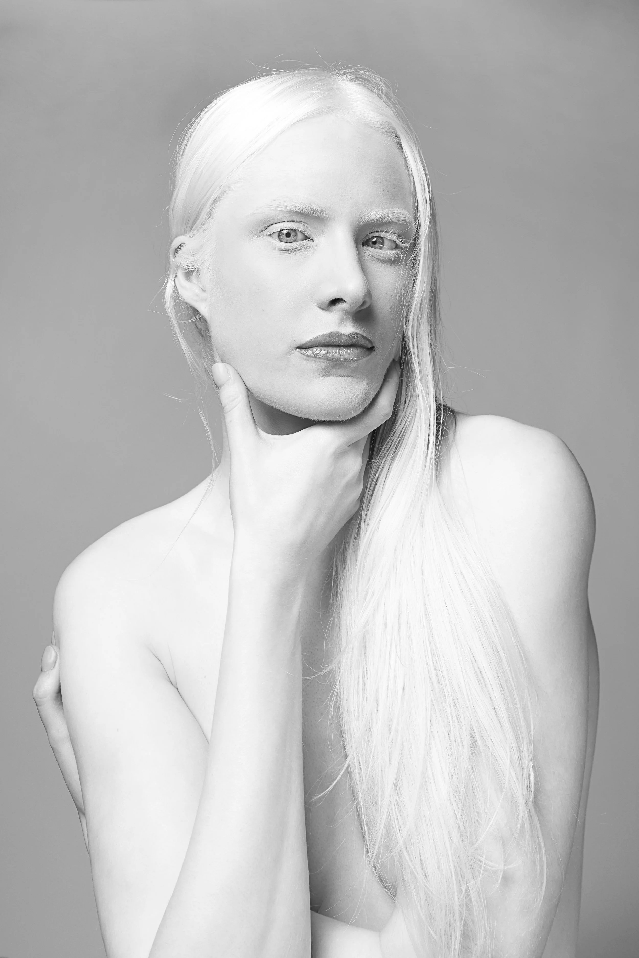 A black and white portrait of a woman with long blonde hair, holding her chin with one hand, looking directly at the camera, with a neutral background.