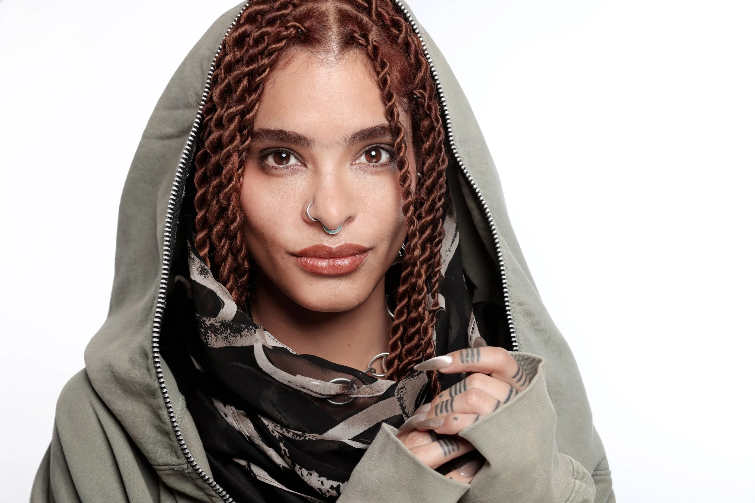 A young woman with curly brown hair and brown eyes wearing a gray hoodie, a black and white patterned shirt, and several facial piercings, including a septum and nose ring, with a subtle smile and holding her hand near her face against a plain white 