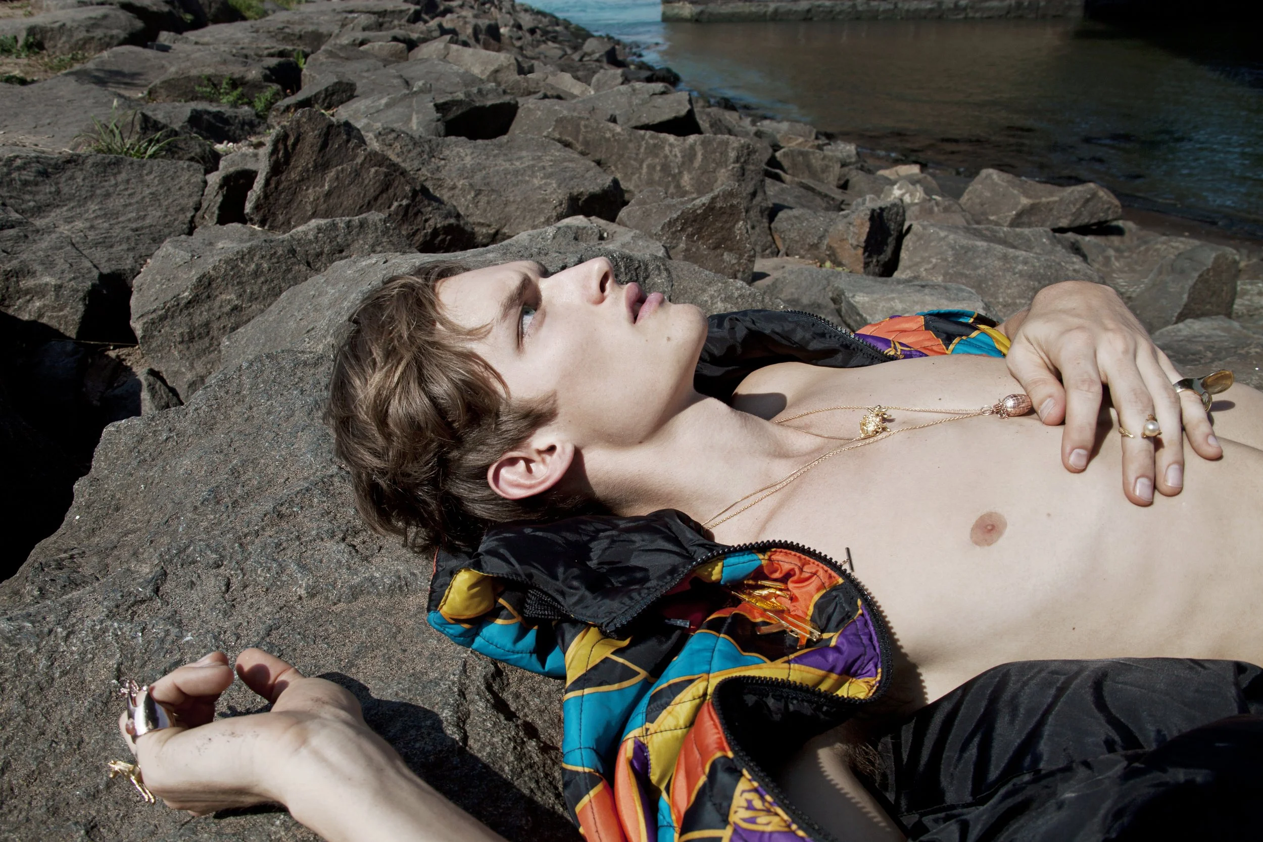 A shirtless man with brown hair lies on a rocky riverbank, wearing a colorful jacket and jewelry, with his hand held by another person.