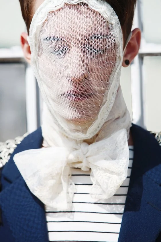 Person wearing a lace face covering with a bow around the neck, dressed in a striped shirt and dark jacket.