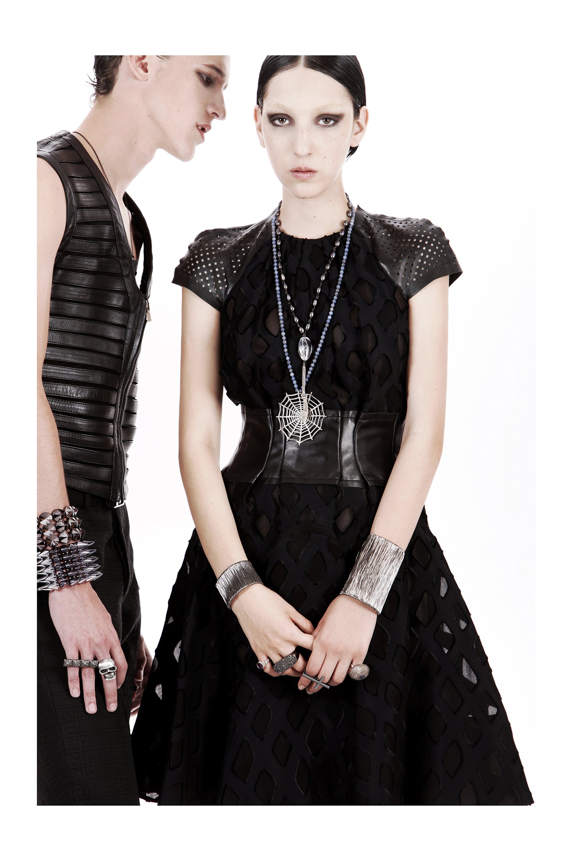 Two women dressed in black with leather and metallic accessories, including jewelry and belts, standing against a white background.