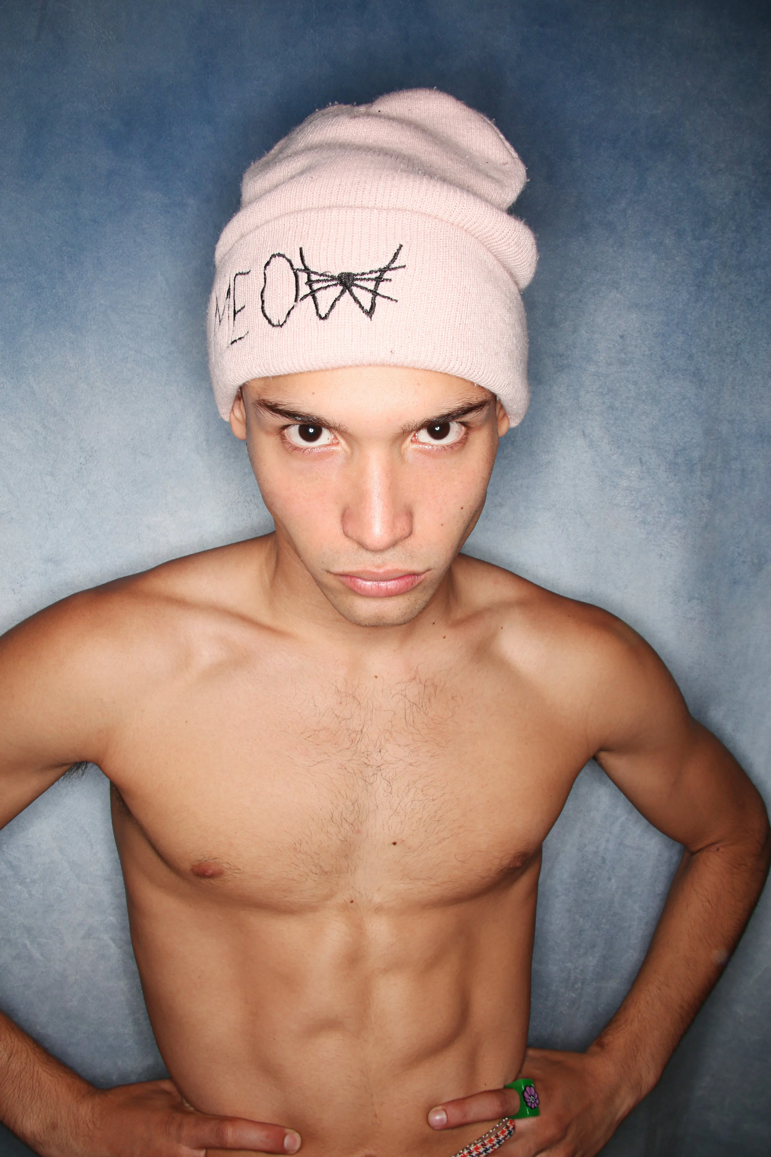 A topless young man with dark hair and brown eyes, wearing a pink beanie with cartoon cat face embroidery, lying on a gray background, looking directly at the camera with hands on hips.
