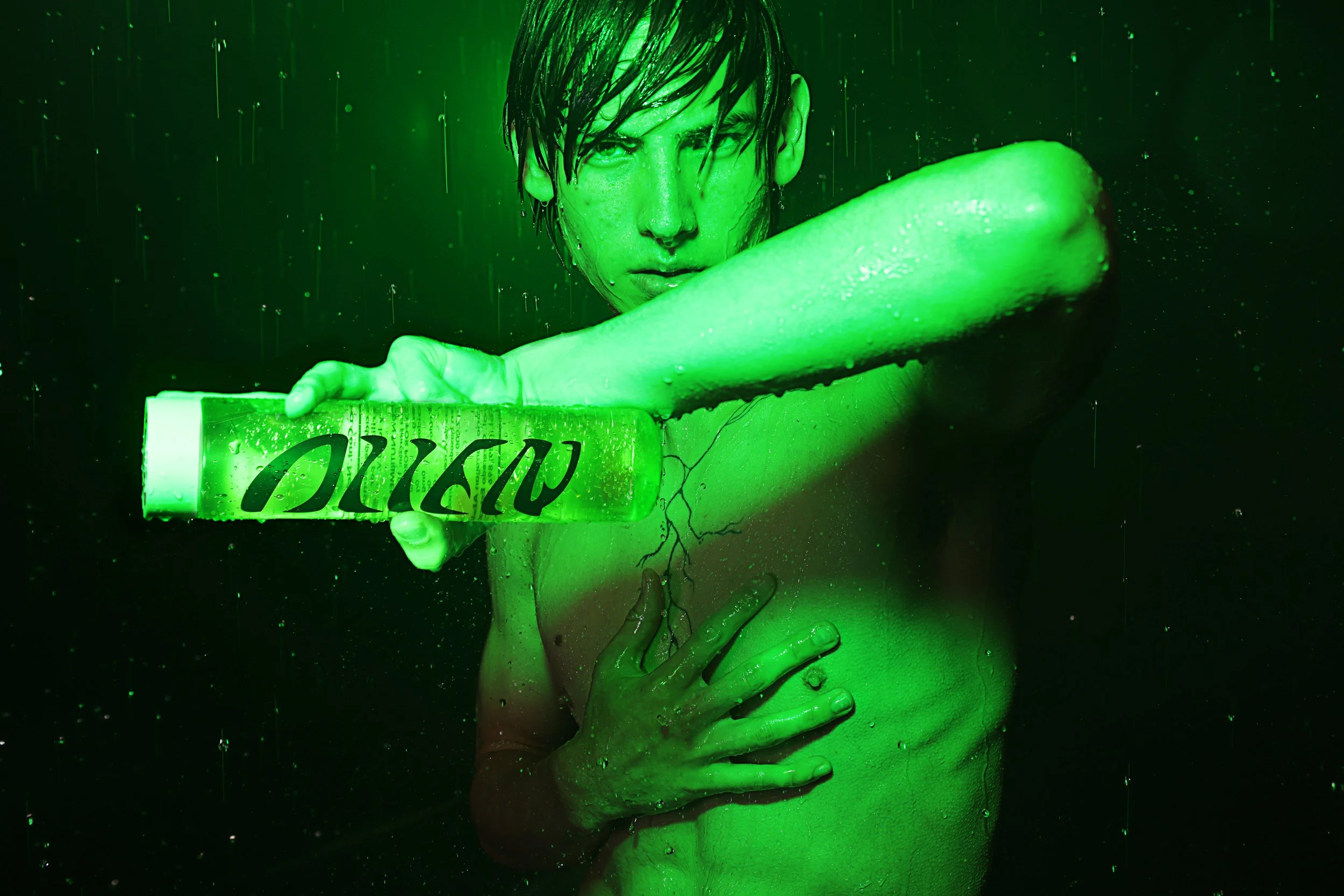 A person with wet hair and a serious expression holding a green bar of soap in front of a green-lit background, with spray and droplets of water visible.