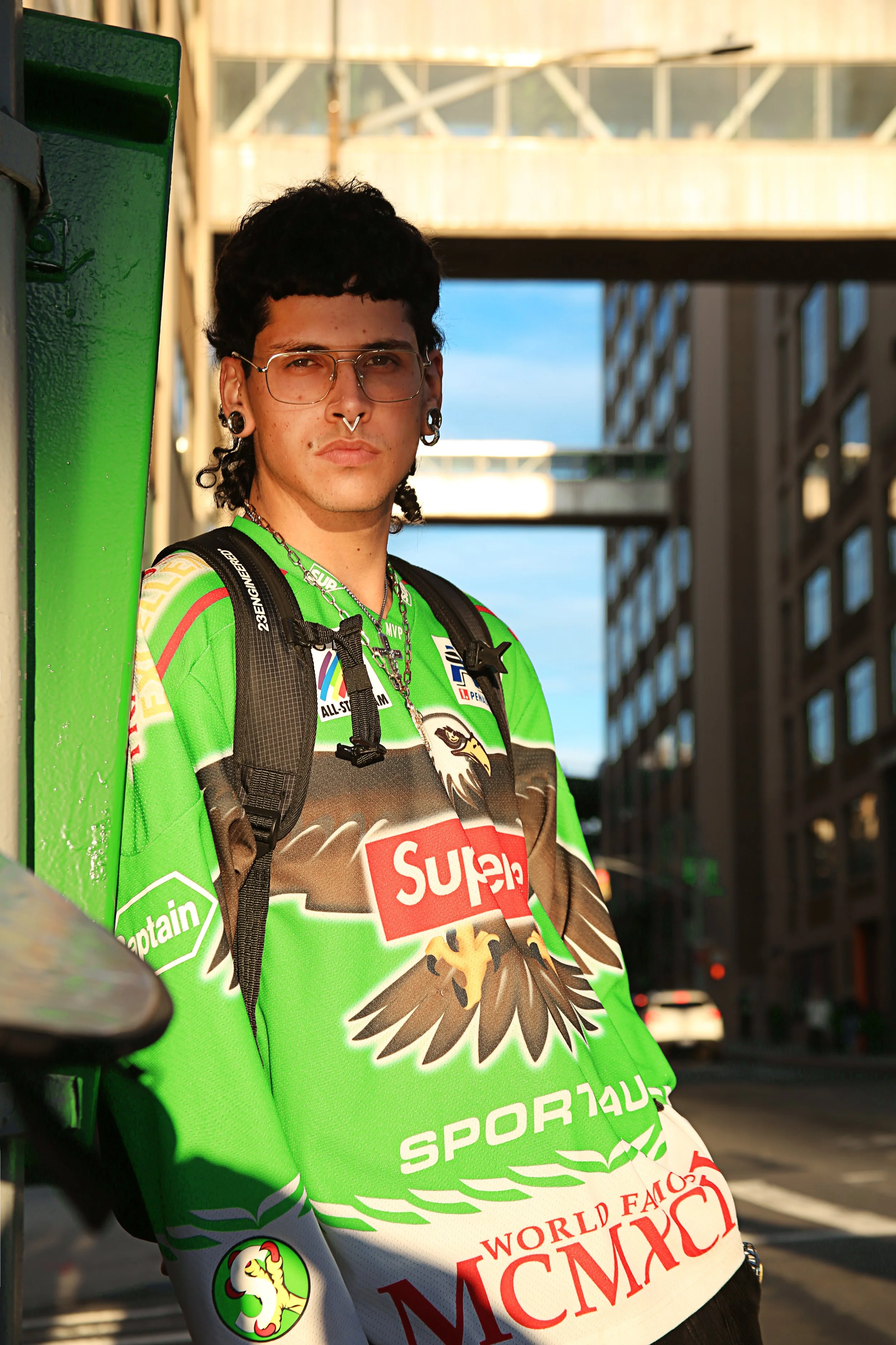 A young man with dark, curly hair, glasses, and extensive earrings, standing outdoors against a city backdrop with a building and sky in the background. He's wearing a bright green sports jersey with logos, a backpack, and a chain necklace, looking d