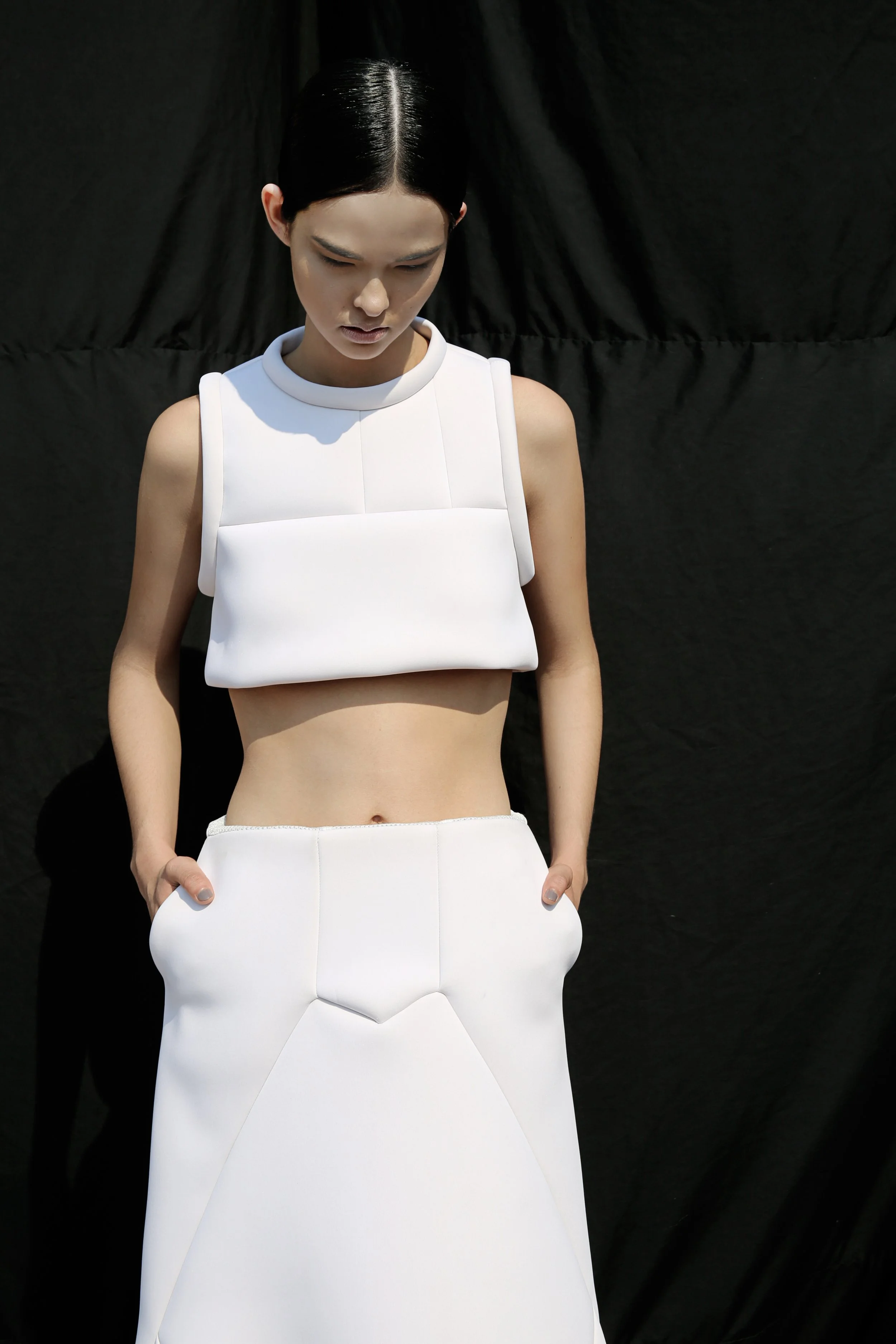A woman dressed in a minimalist white outfit with a crop top and matching trousers, standing against a black backdrop, looking downward with her hands in her pockets.