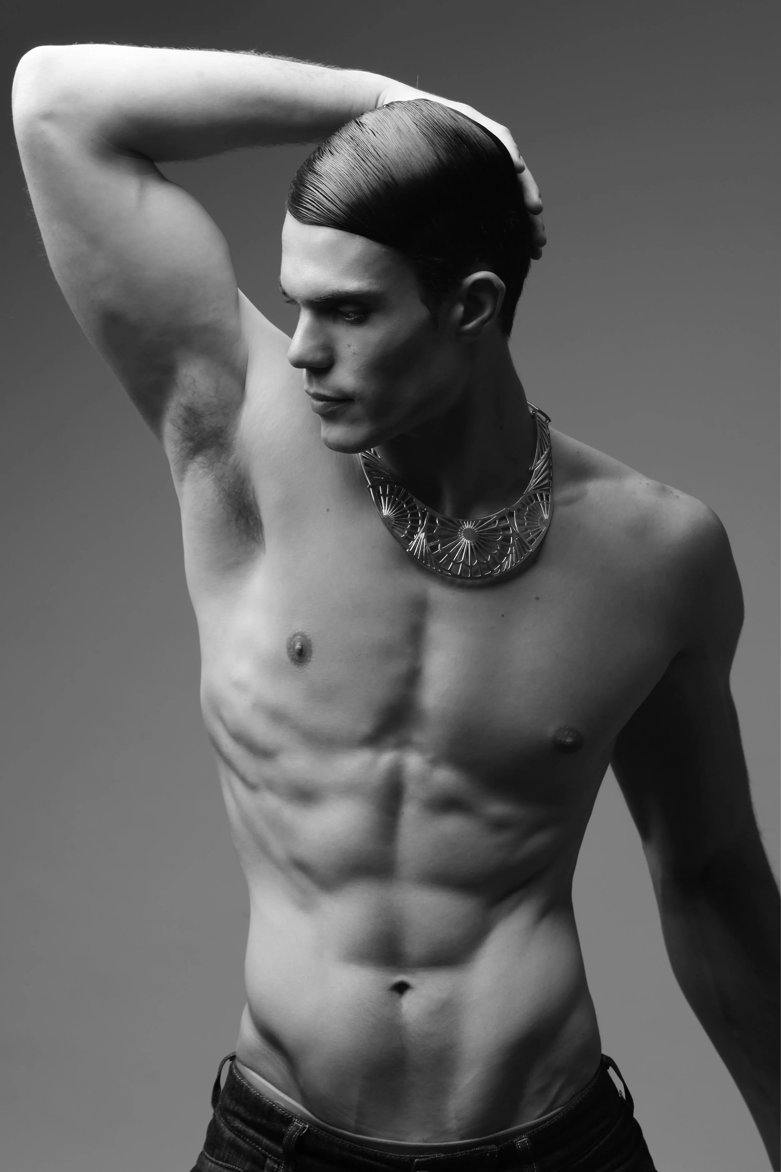 A black and white photo of a shirtless man with a muscular build, slicked-back hair, and an elaborate necklace, posing with one hand on his head against a plain background.