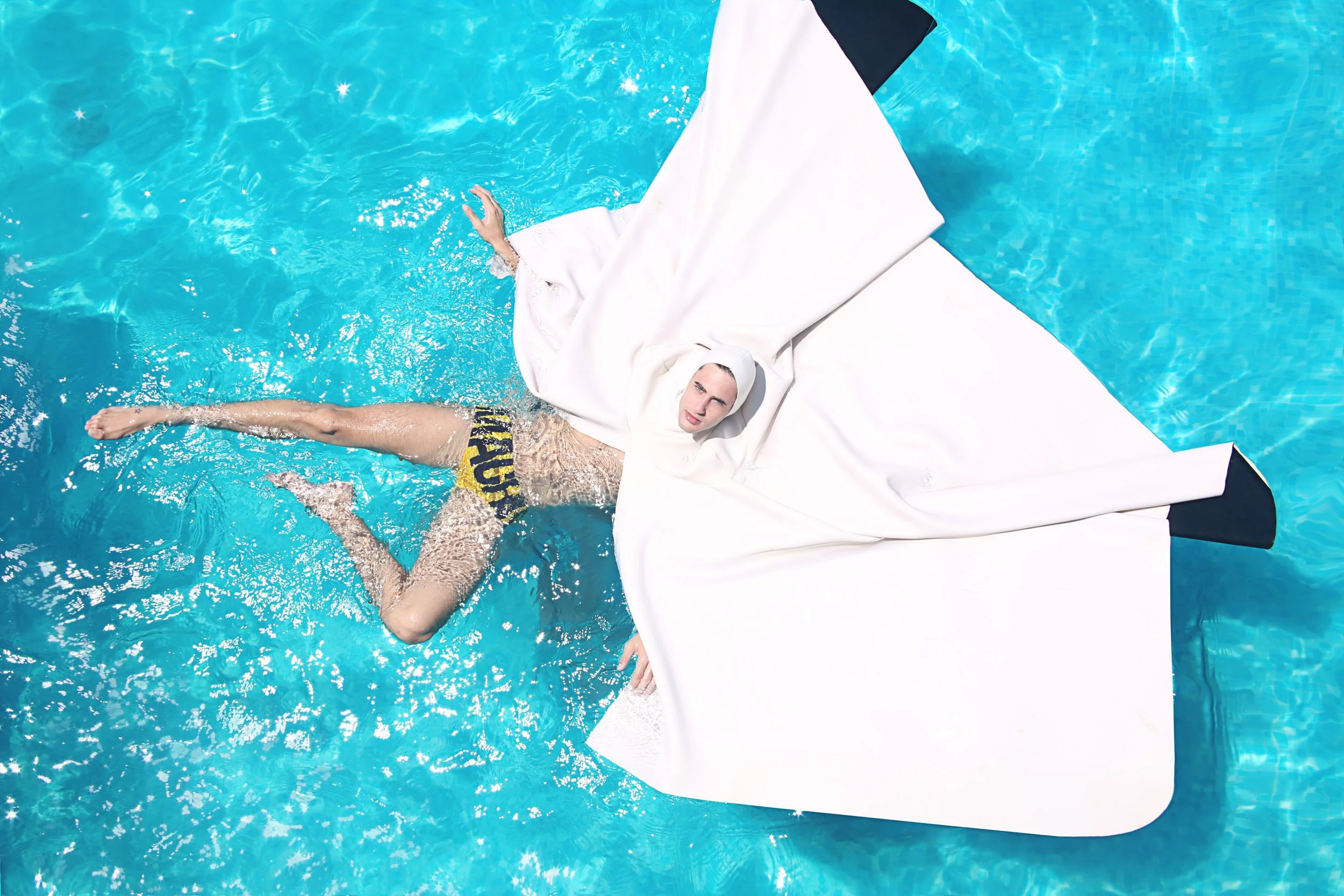 Person lying in a swimming pool on an inflatable float, wearing black and yellow swim trunks, with one arm resting on the float and the other arm submerged in the water.