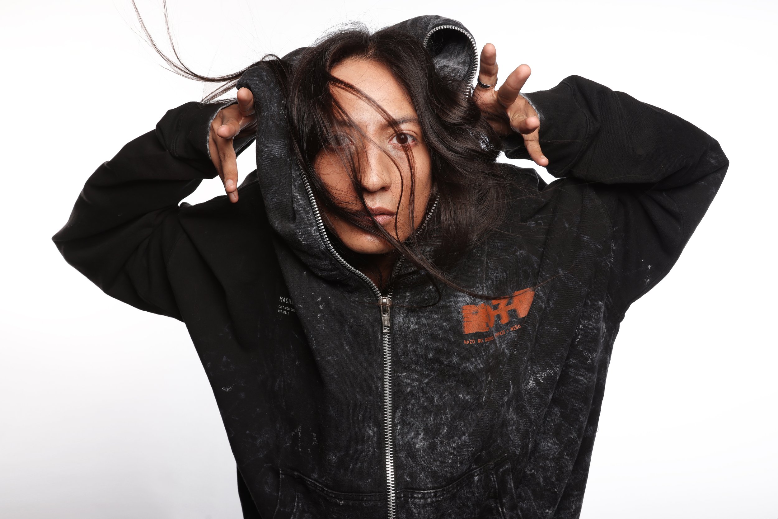A person with long dark hair, wearing a black hoodie, holding the hood with both hands, looking directly at the camera, with a white background.