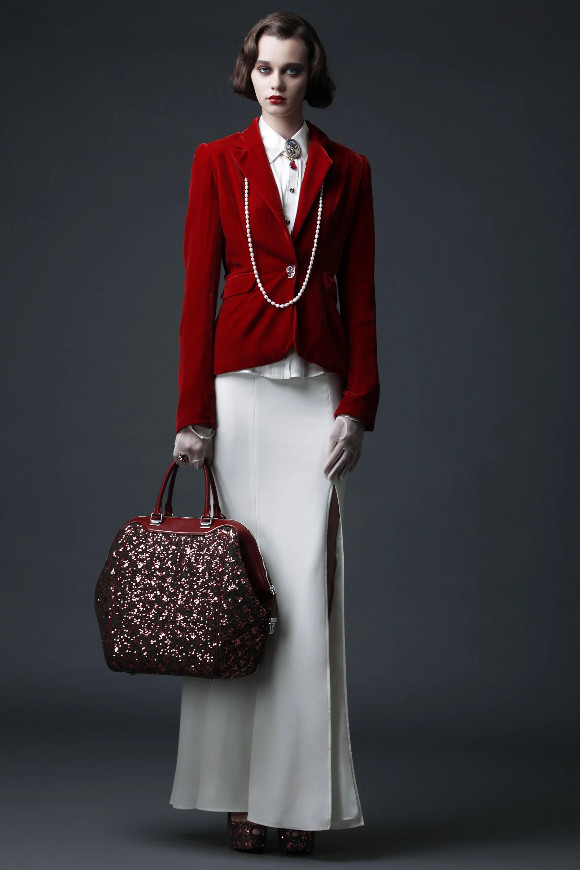 A woman with short brown hair wearing a red velvet blazer, white blouse, and long white skirt with a slit, standing against a dark background. She accessorizes with a long pearl necklace, brooch, rings, and holds a sequined red and black handbag. She