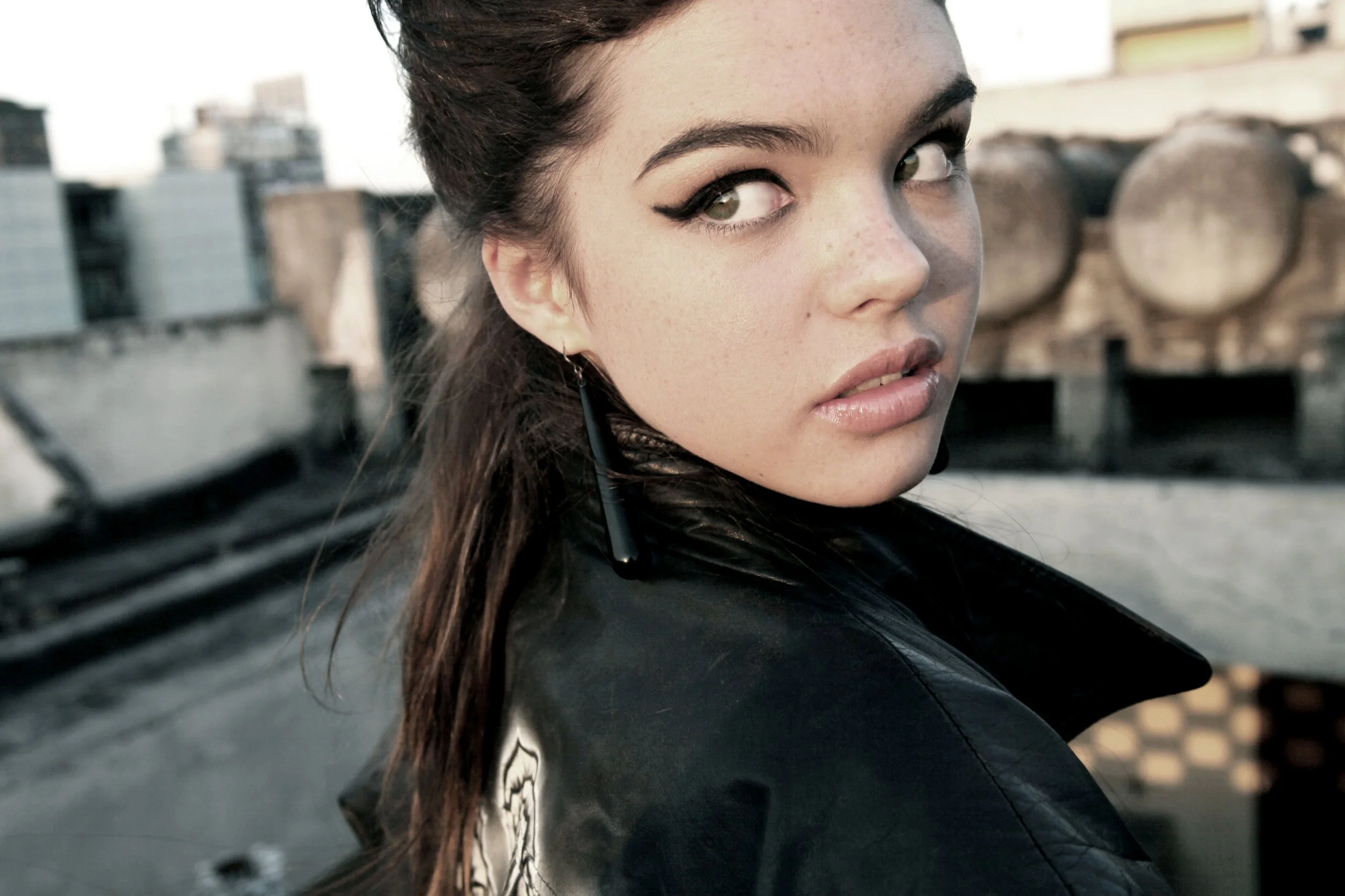 Close-up of a woman with dark hair, winged eyeliner, and earrings, looking over her shoulder on a rooftop with a cityscape background.
