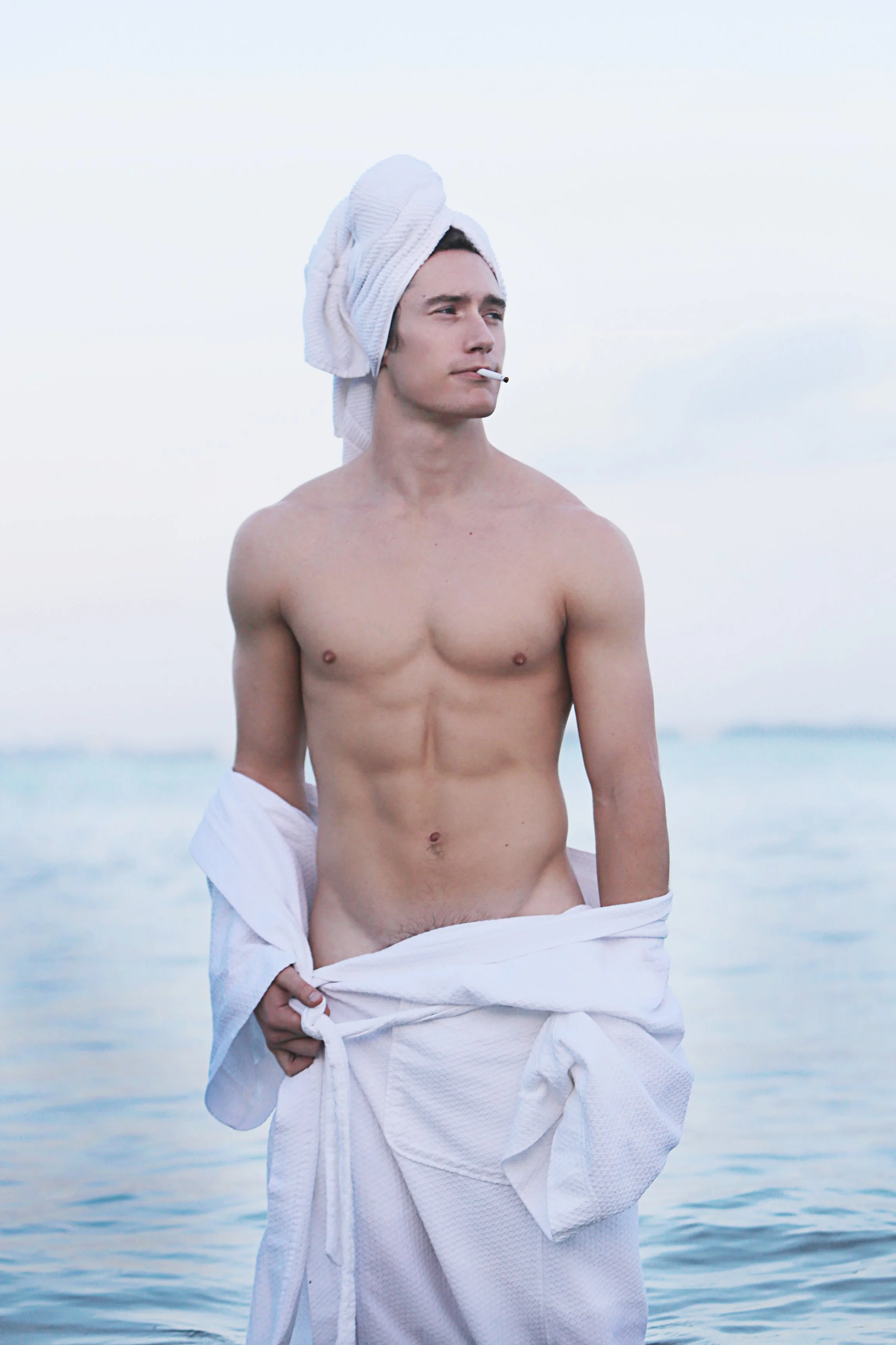 A shirtless young man with a towel wrapped around his head, holding a white bathrobe at the waist, standing by the beach with the ocean in the background, smoking a cigarette.