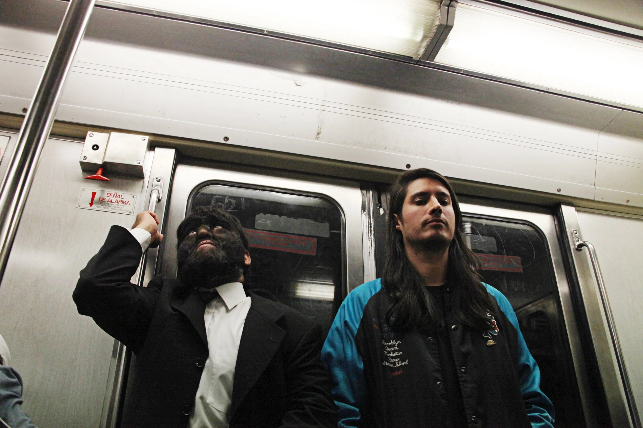 Three people standing inside a subway train. The person on the left is a man dressed in a black suit, the person in the middle is a woman with long dark hair wearing a black jacket with various words on it, and the person on the right is a man with l