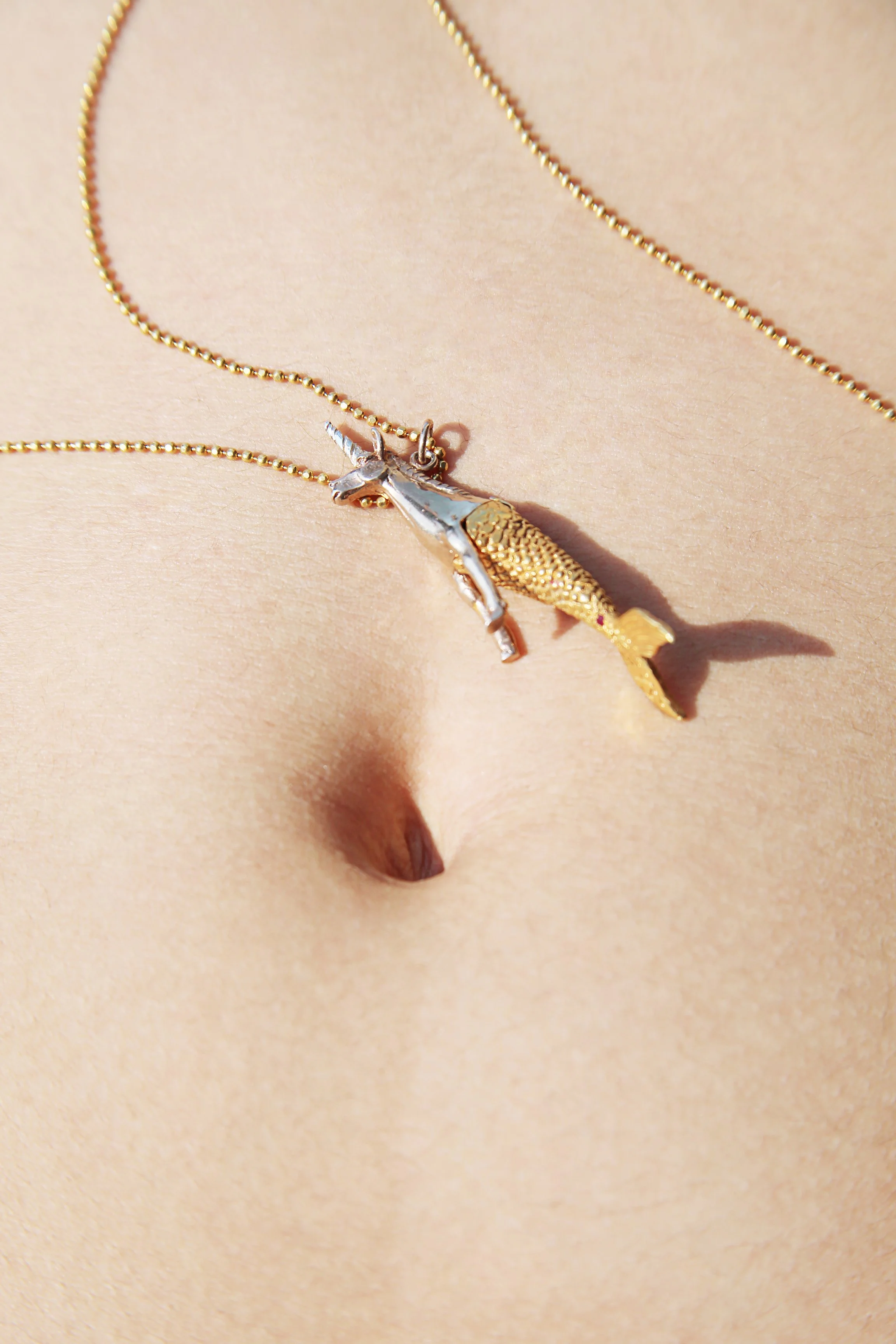A close-up of a person's belly button with a gold necklace featuring a unicorn and a fish pendant resting on their skin.