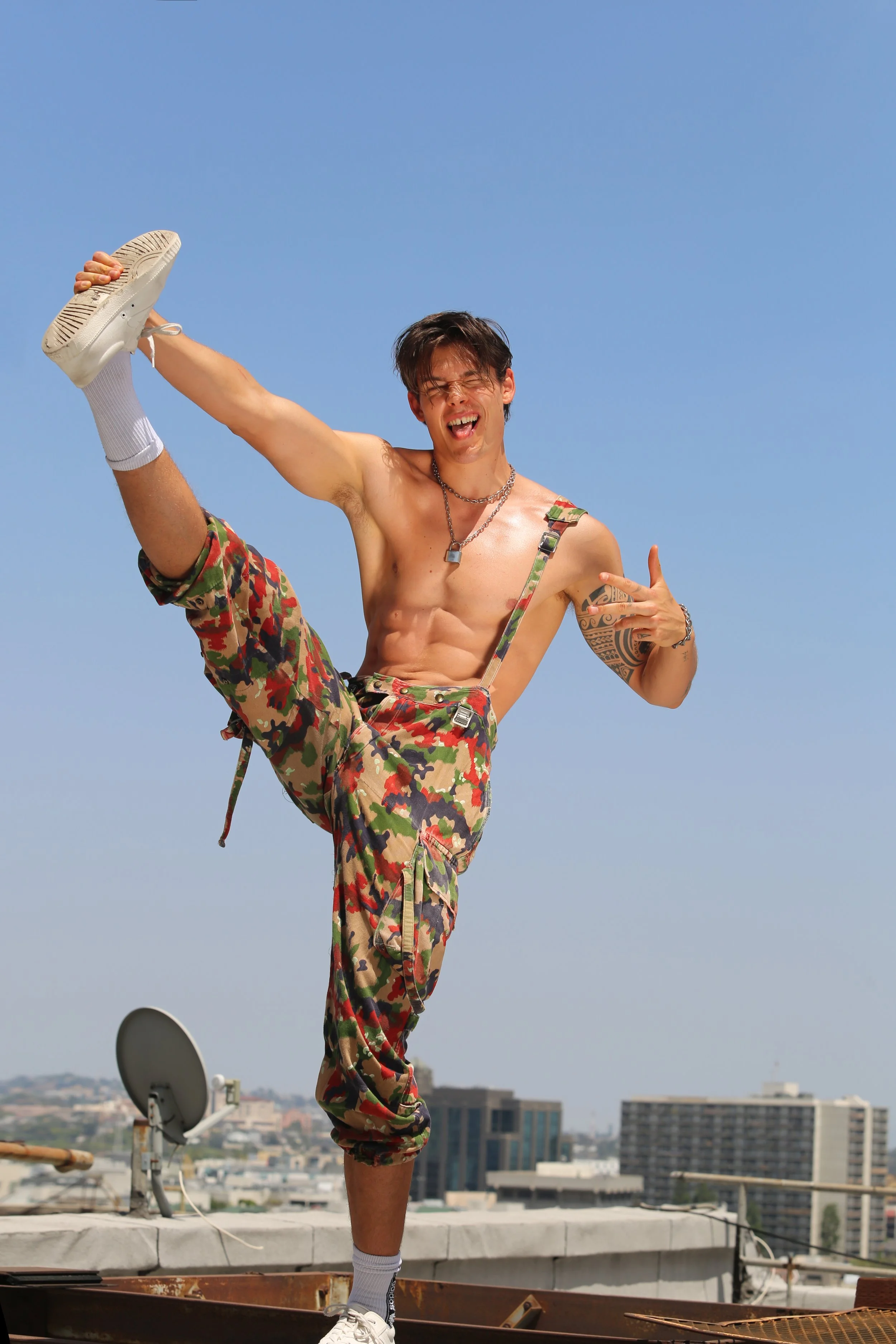 A young man with dark hair, shirtless, wearing camouflage shorts, white sneakers, and socks, striking a high kick on a rooftop under a clear blue sky.