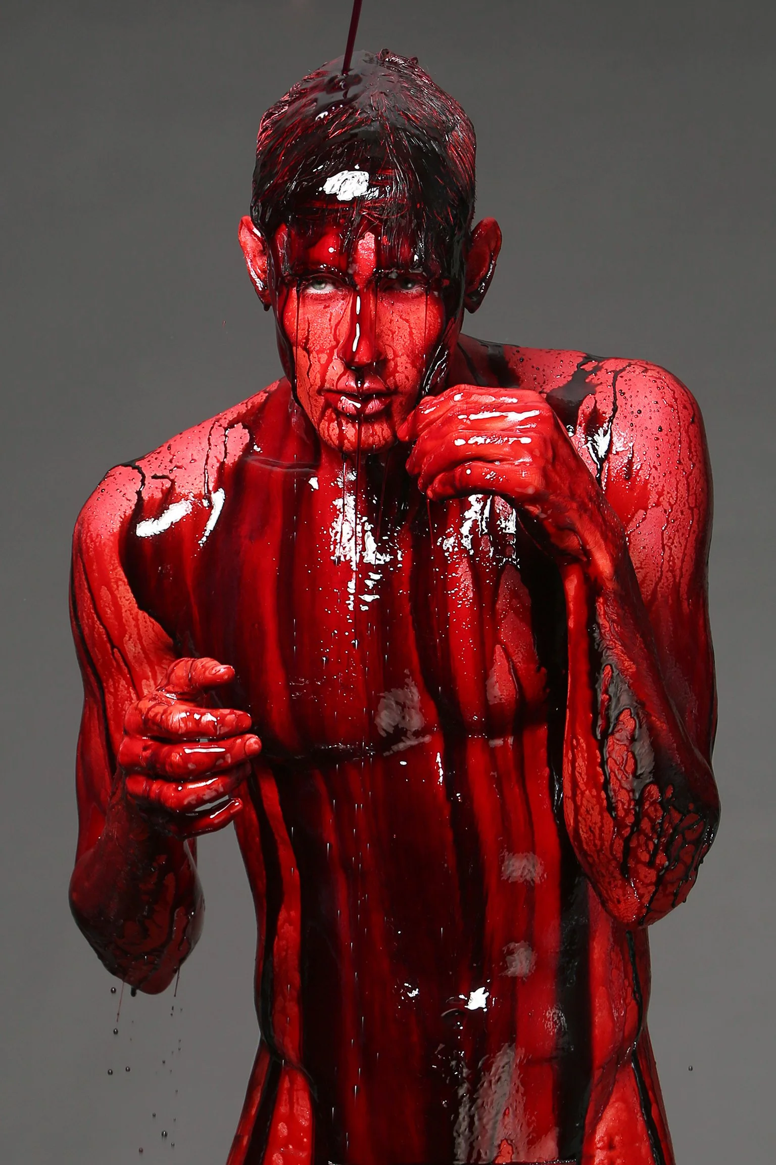 Person covered in red and black paint, with paint dripping down their face and body, holding their fist close to their mouth against a gray background.