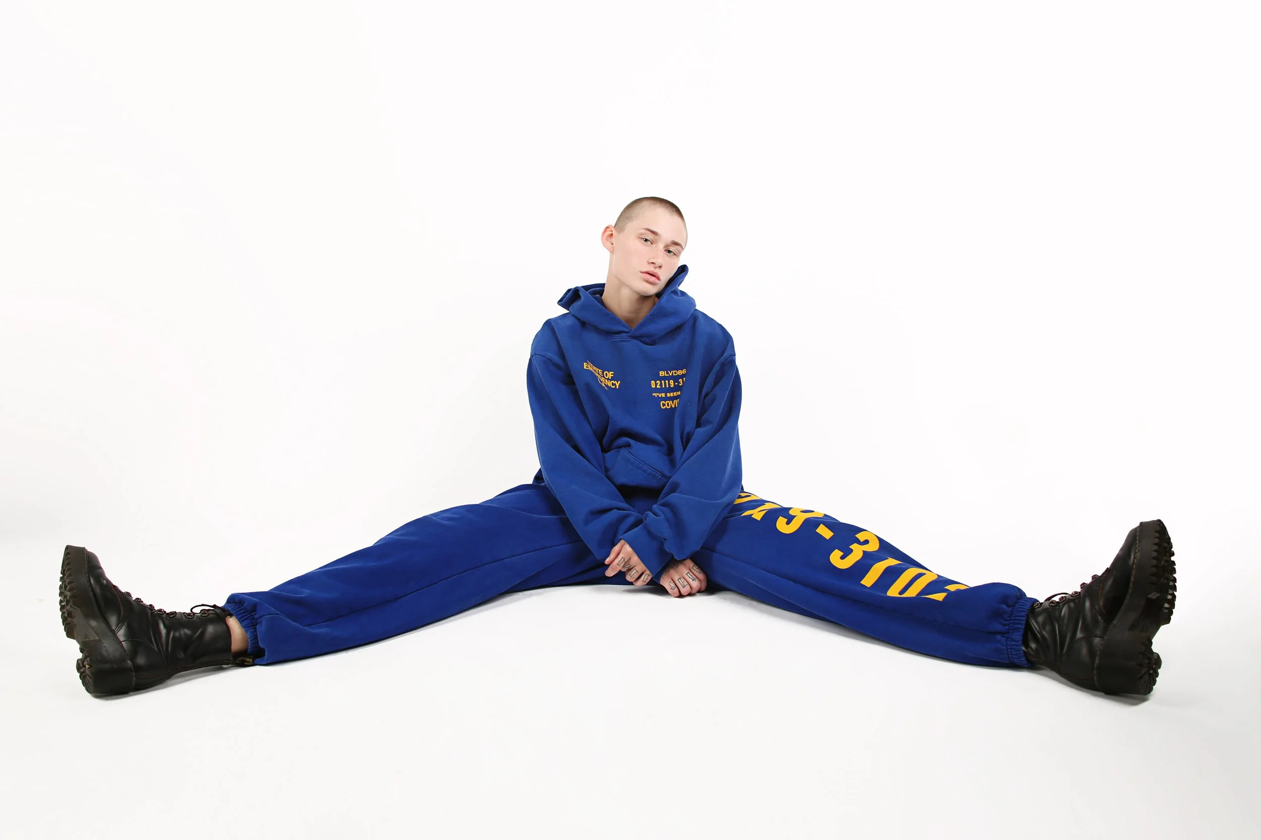 A young person with a shaved head sitting on the floor with legs spread wide. They are wearing a blue hoodie and matching blue sweatpants with yellow text on the left leg. They are also wearing black combat boots. The background is plain white.
