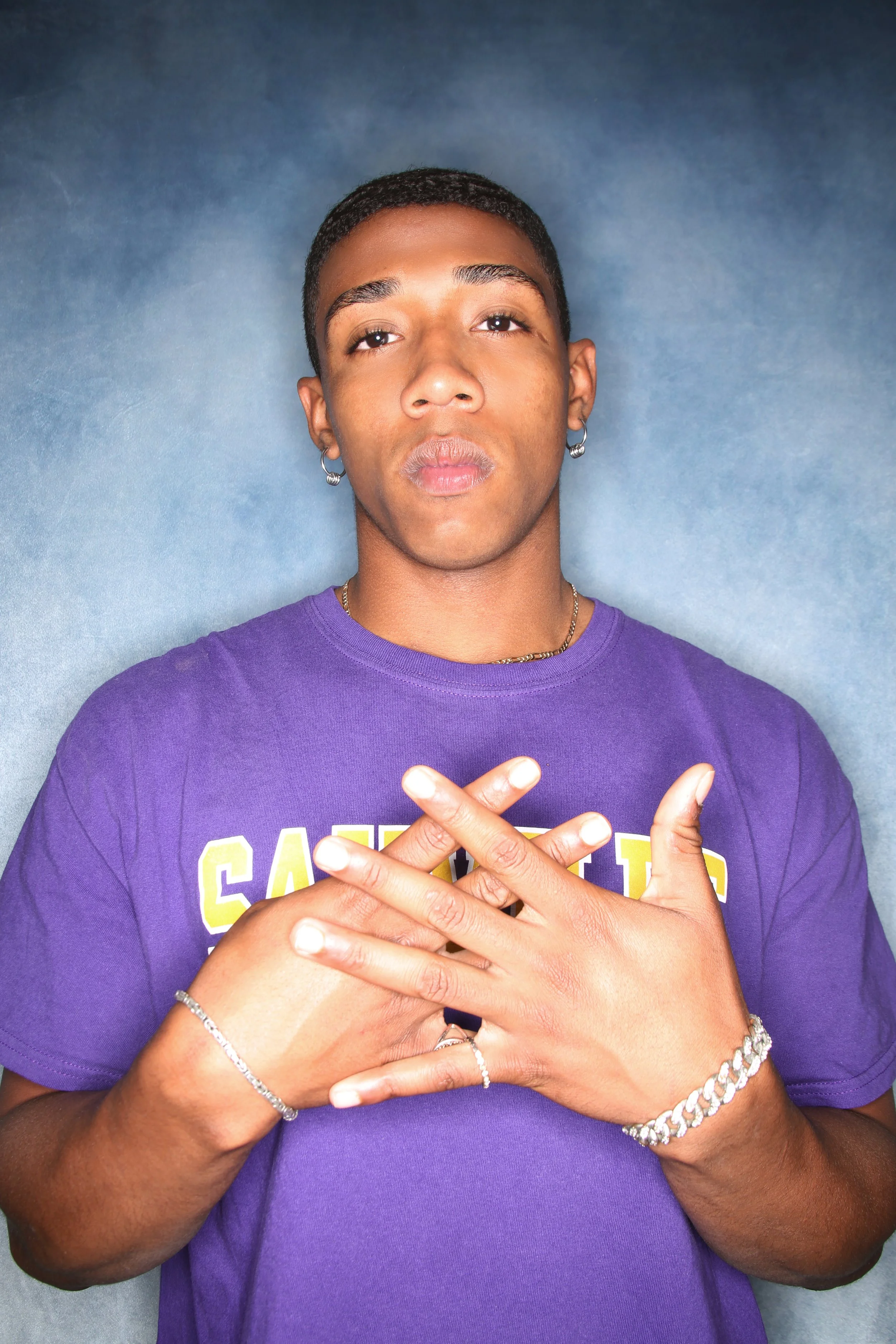 A person wearing a purple shirt with yellow and white text, has their hands crossed over their chest, and is wearing jewelry including rings, bracelets, earrings, and a necklace, against a dark blue background.