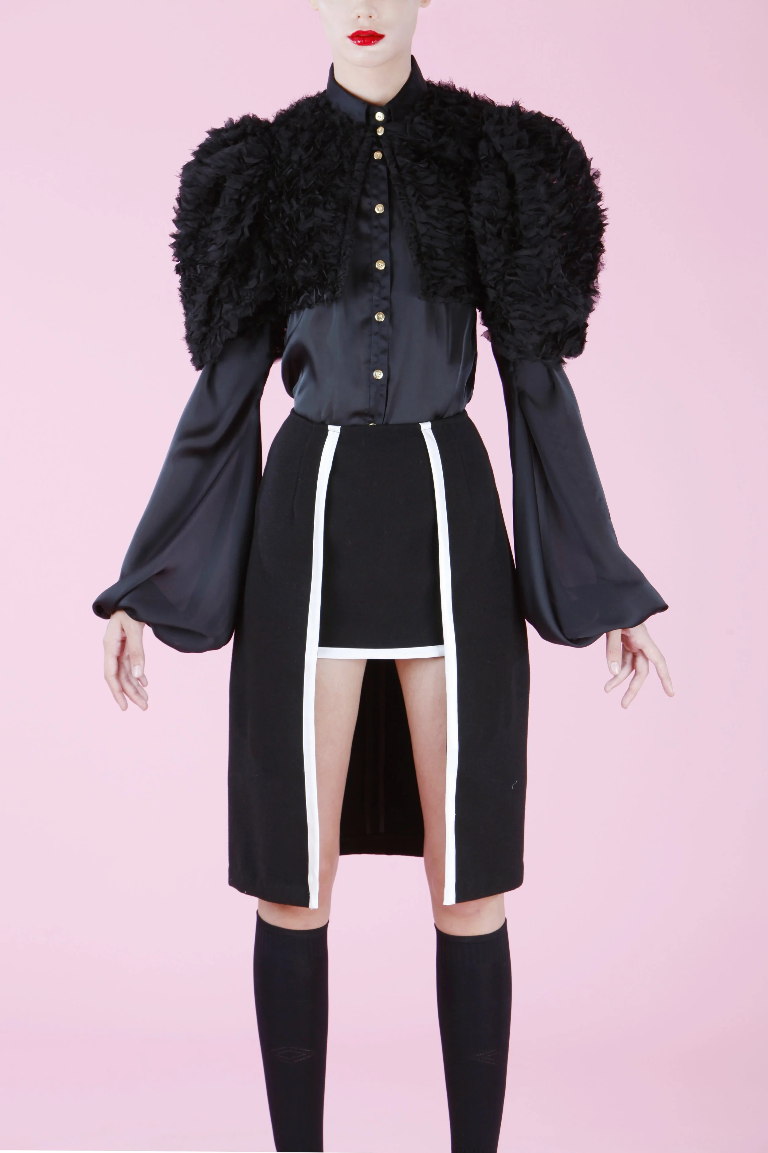 A woman with red lipstick wearing a black shirt with puffy, feather-like black shoulder accents, a black skirt with white vertical stripes, and black thigh-high socks, posed against a pink background.