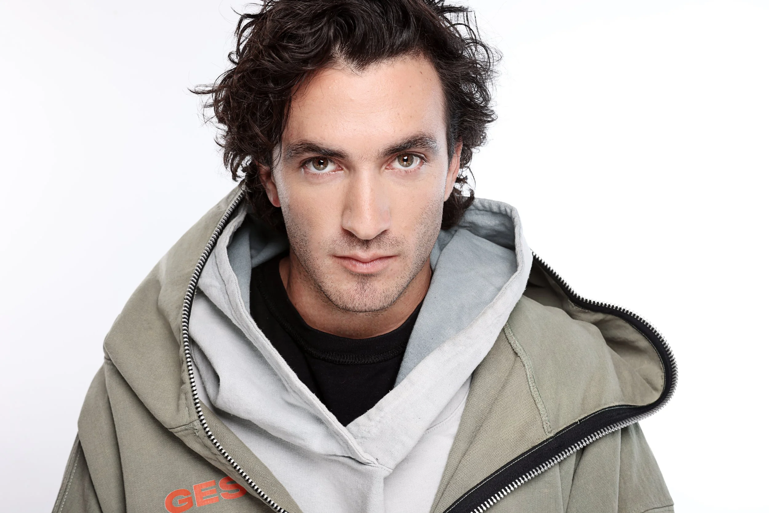 A man with curly dark hair looking directly at the camera, wearing a light gray jacket over a white hoodie.