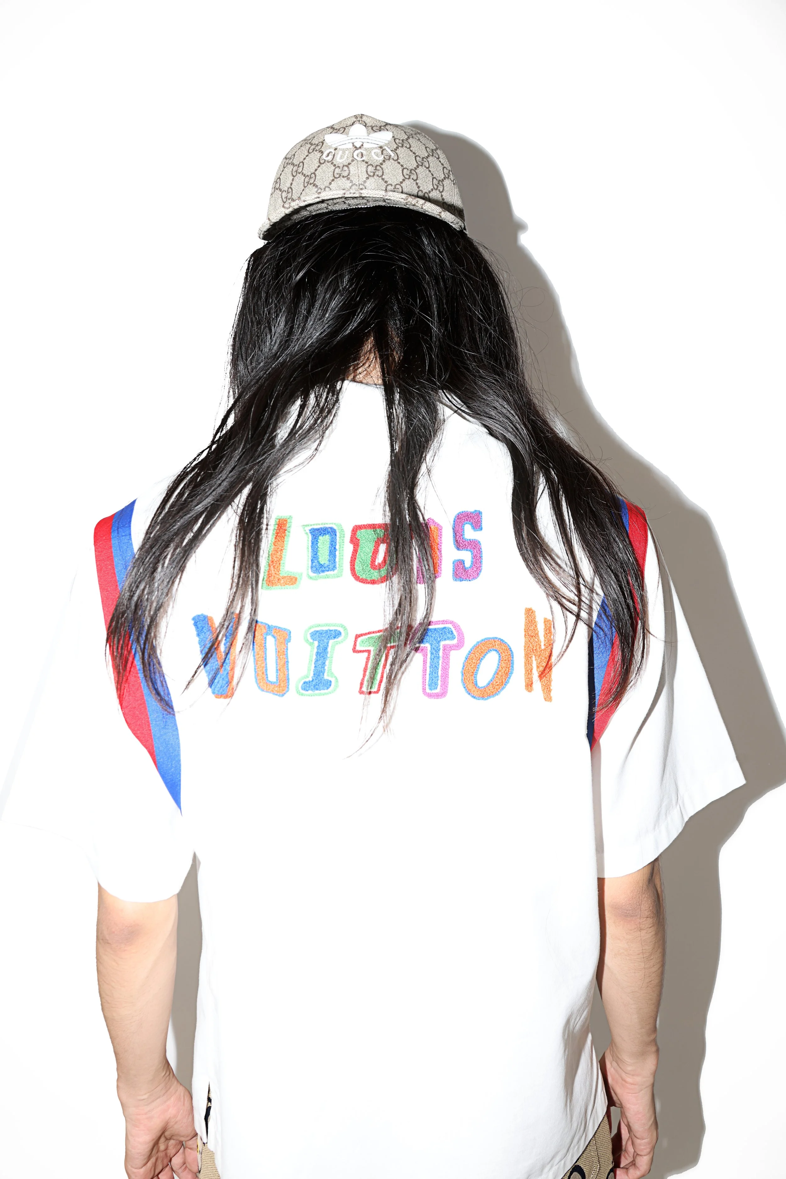 Back of a person with long black hair wearing a white t-shirt with colorful embroidered text and a beige Gucci baseball cap, standing against a plain white wall.