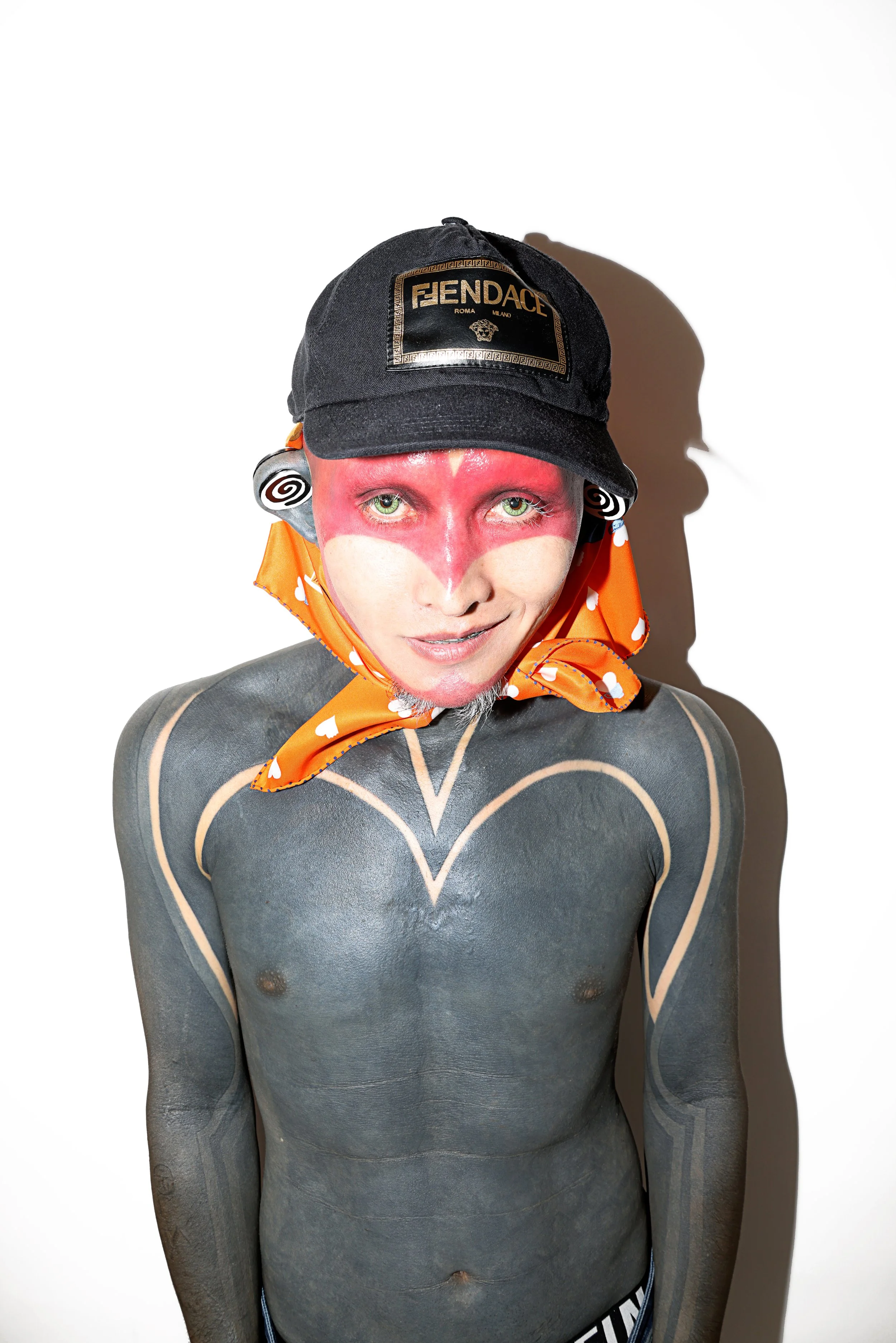 Person with gray skin body paint and body markings, wearing an orange scarf with white hearts, a black Fendi cap, and spiral earrings, standing against a white background.