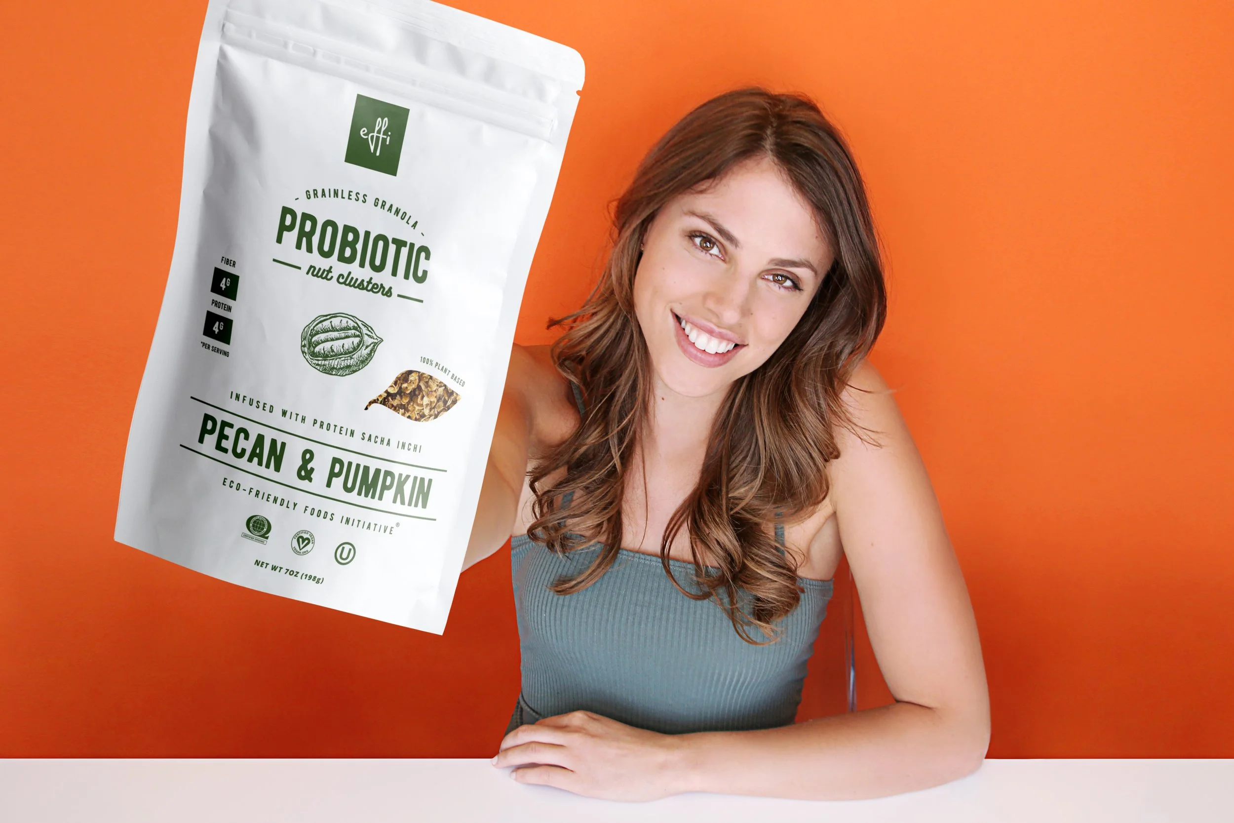 Smiling woman with long brown hair wearing a gray tank top, sitting at a white table against an orange background, with a white bag of Effi probiotic granola featuring green and black text and illustrations in the foreground.