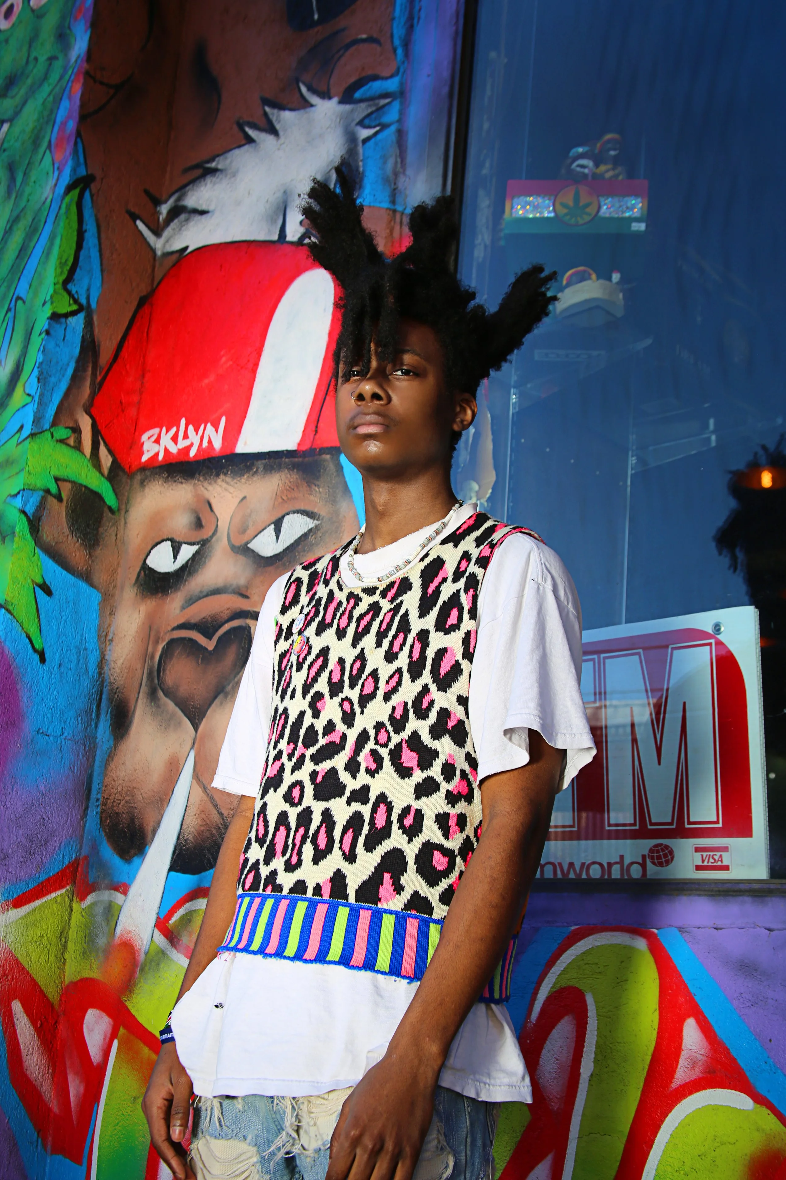 A young man with styled hair stands in front of vibrant street art depicting a child with a hat labeled 'BKLYN' and graffiti-style lettering.