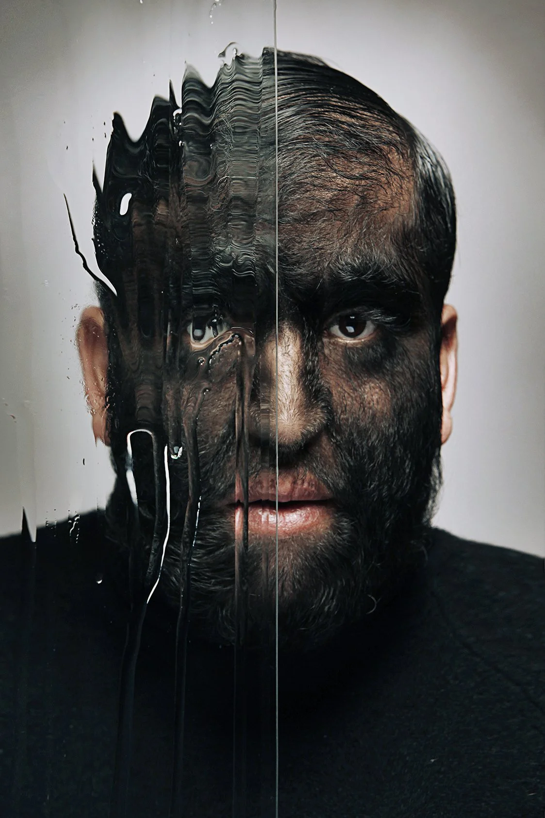 A man with dark hair and a beard, half of his face is mirrored and obscured with black paint, water, or ink on a glass surface.