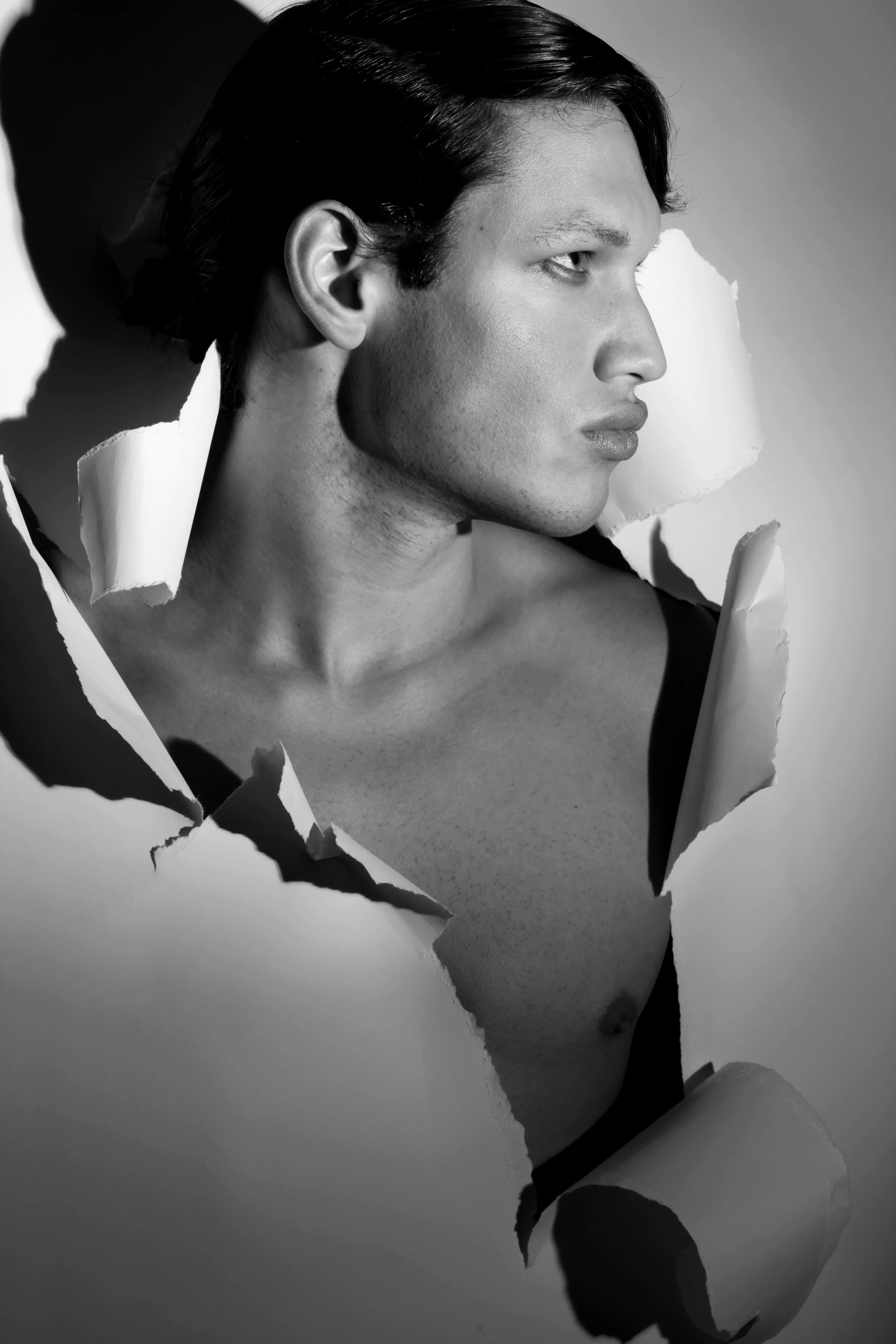 A black and white photo of a young man with dark, slicked-back hair, looking to the right. The image appears to be emerging from torn paper, creating a frame around his face and upper torso.
