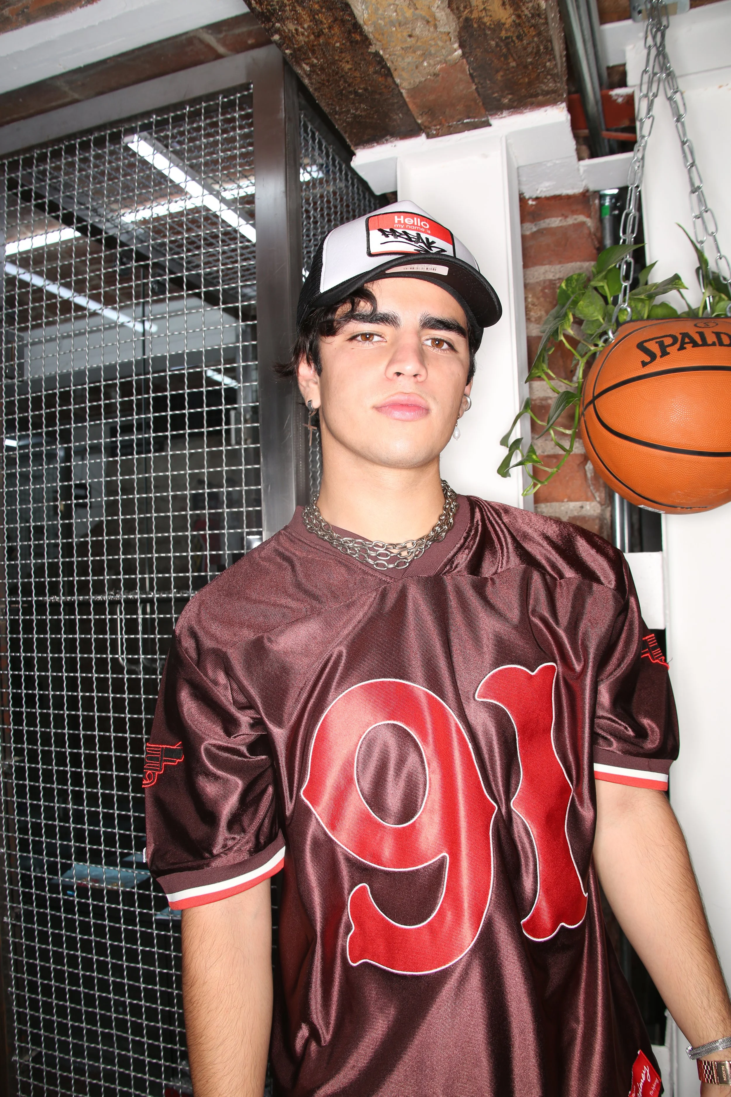 A young man with dark hair, wearing a NFL football jersey with the number 99, a black and white cap with a name tag on it, earrings, jewelry, standing in an industrial-style room with exposed brick wall, chain hanging from the ceiling, a basketball, 