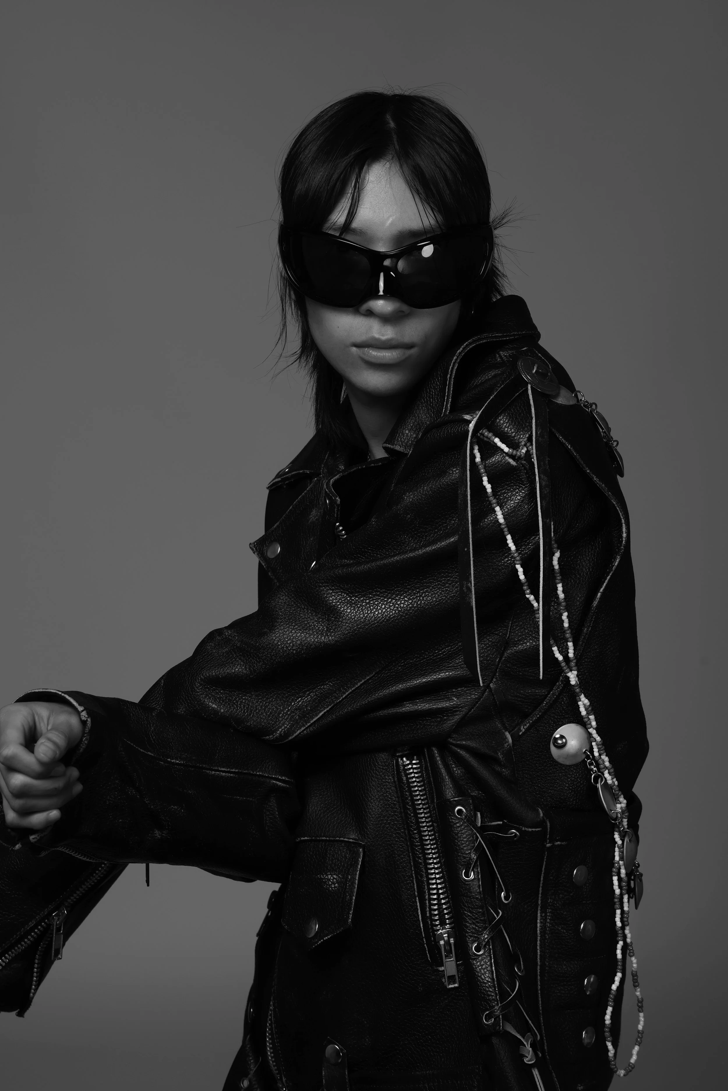 Black and white photo of a person with shoulder-length dark hair wearing large sunglasses, a leather jacket with zippers and metal details, and decorated with beads and accessories.