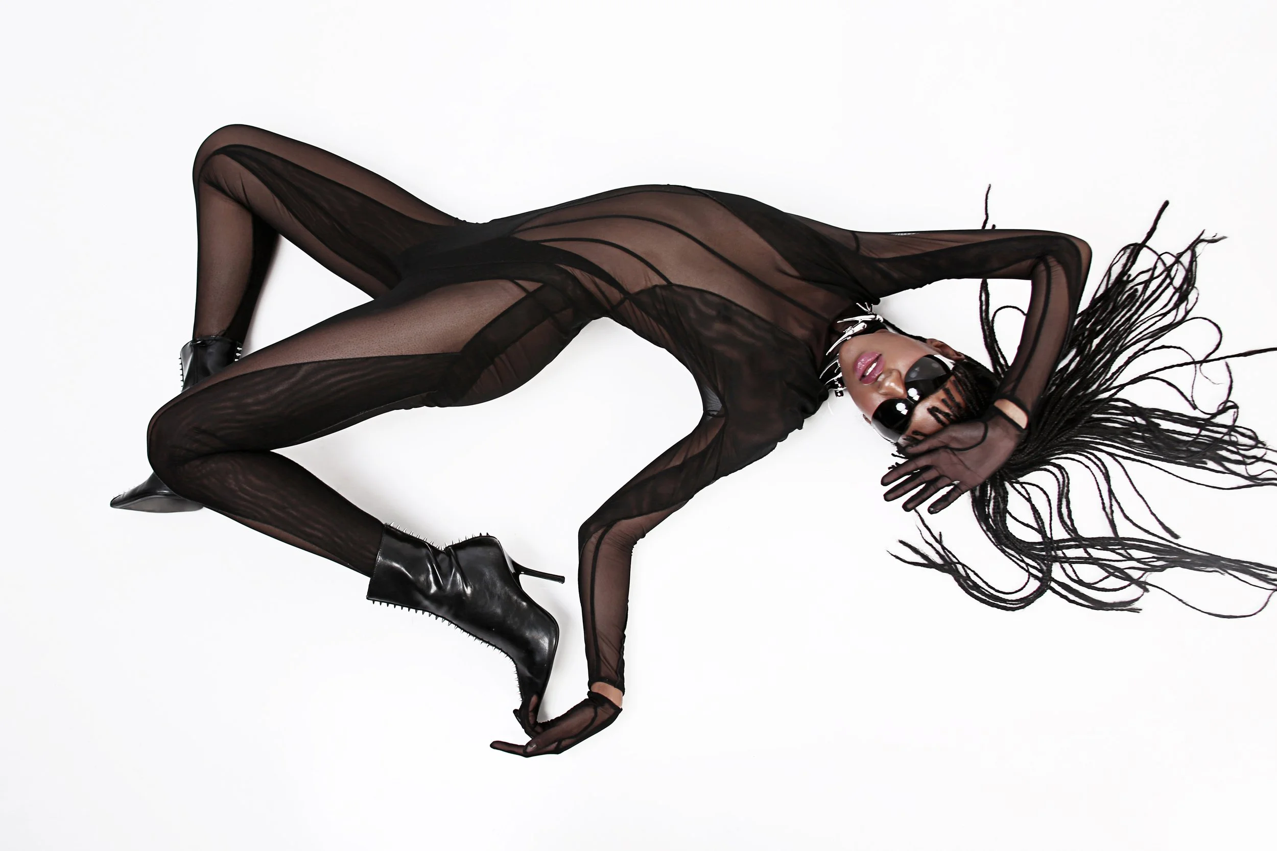 A woman with long braided hair lying on a white background, wearing a sheer black outfit and black high-heeled boots, with sunglasses and jewelry.