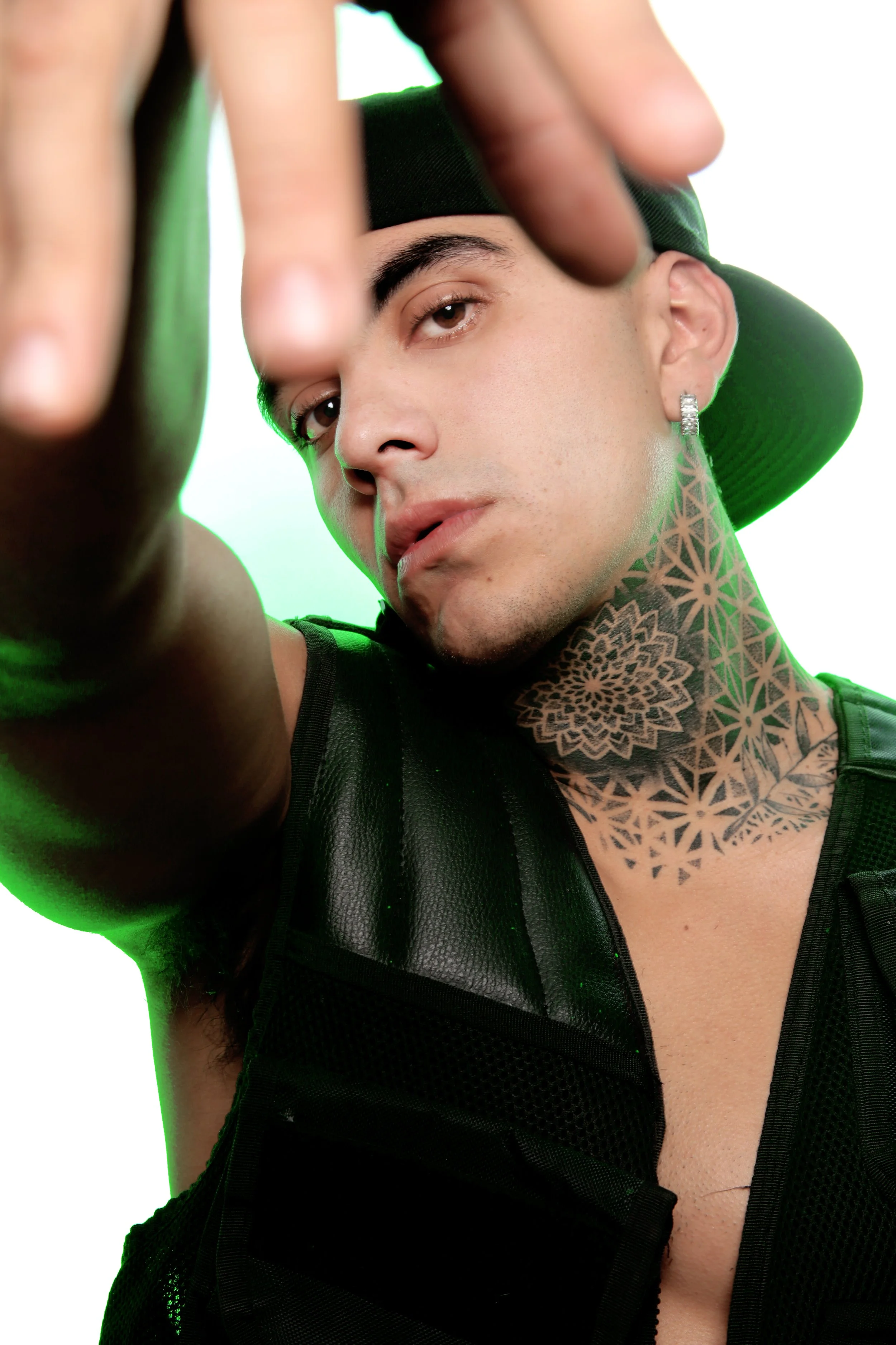 Close-up of a young man with tattoos on his neck, wearing a black hat and earrings, reaching towards the camera with a serious expression.