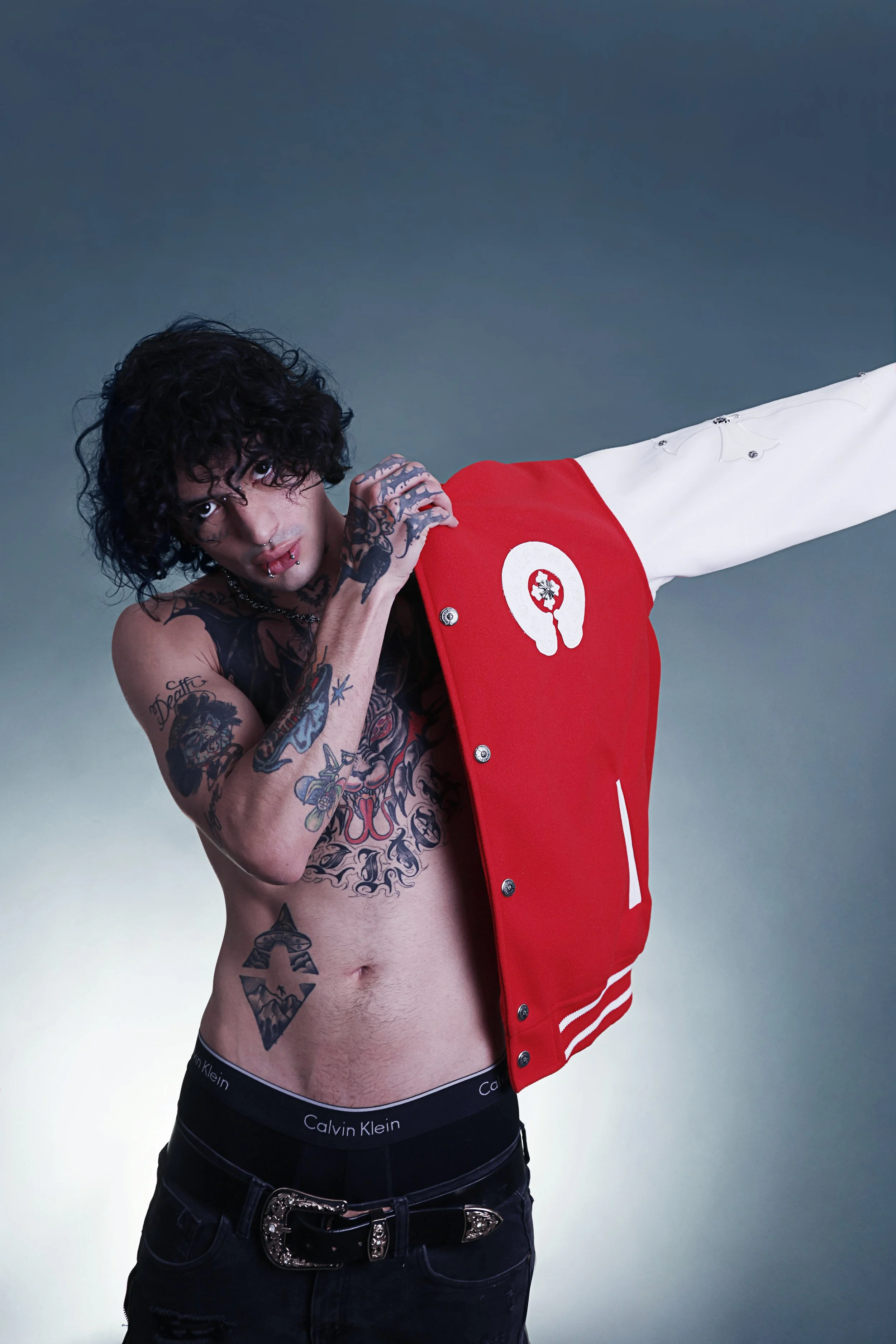 A shirtless man with tattoos, black curly hair, and piercings is holding a red and white jacket. He is wearing black Calvin Klein underwear, black jeans with an embellished belt, and is posing against a neutral background.